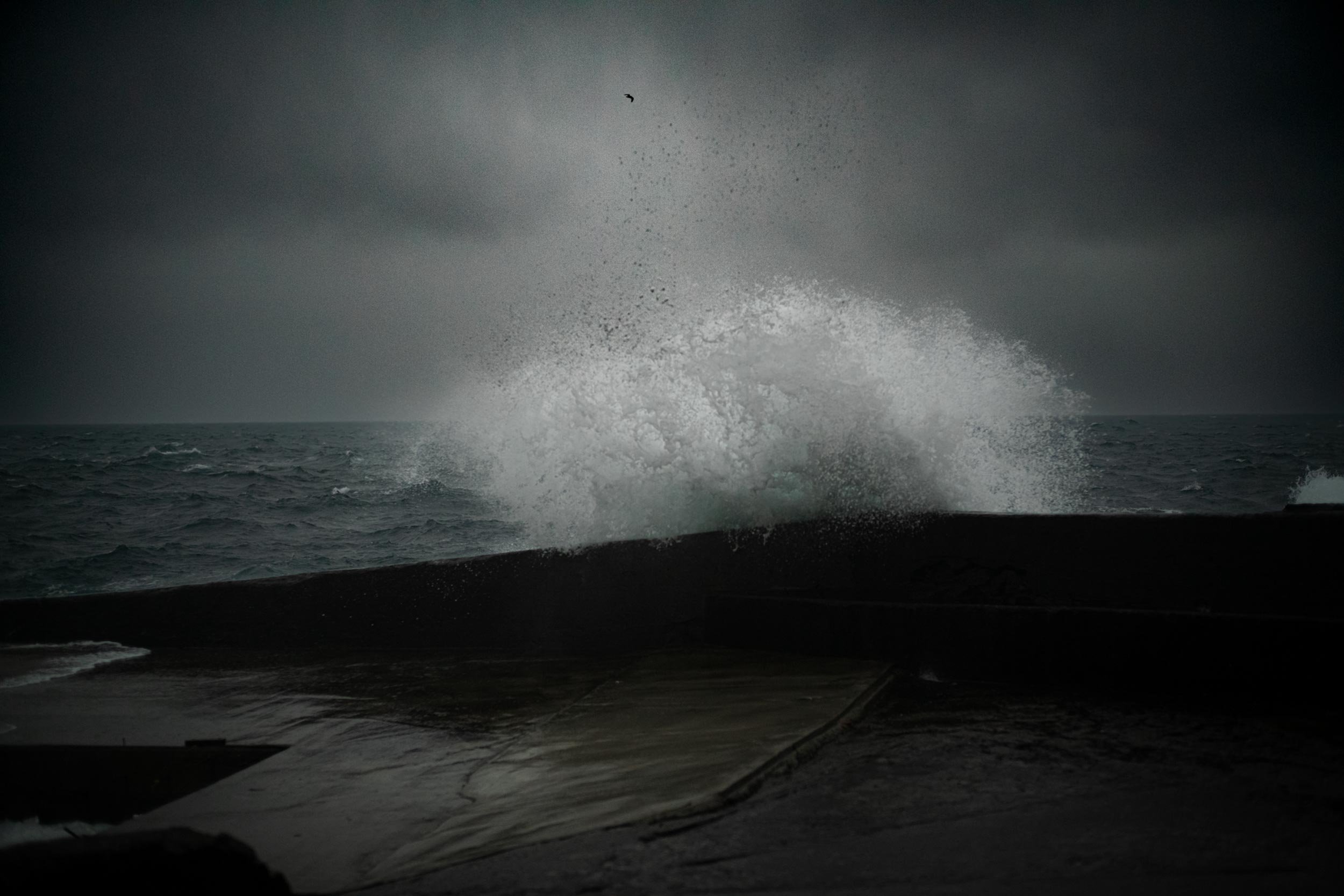 Grosse welle spritzt ueber eine dunkle hafenmauer bei bedecktem himmel, faeroer