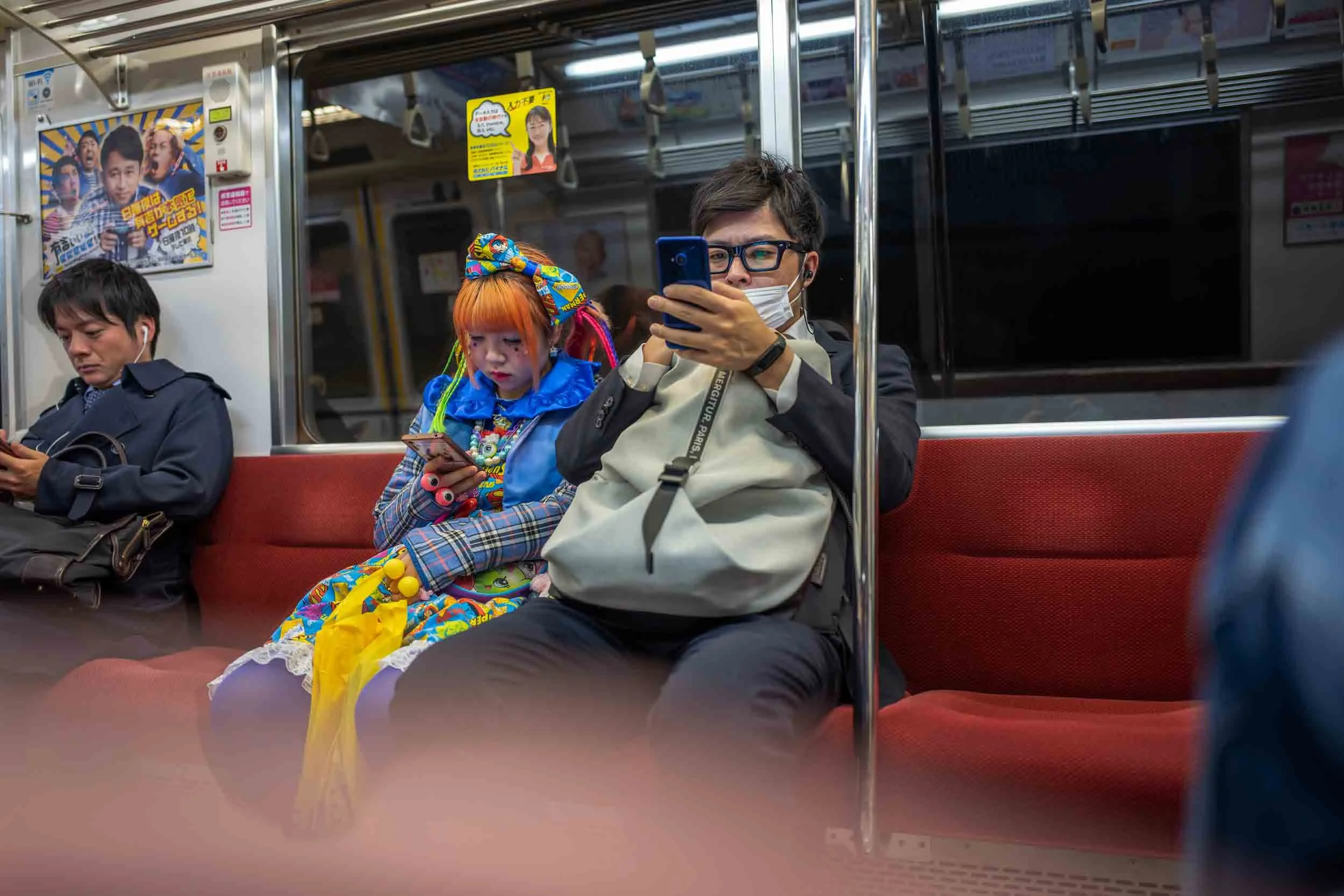 Unterschiedliche Menschen sind in ihre Smartphones vertieft, darunter eine bunte, auffällig gekleidete Person. Der Moment zeigt den Kontrast zwischen traditioneller Zurückhaltung und moderner Individualität.