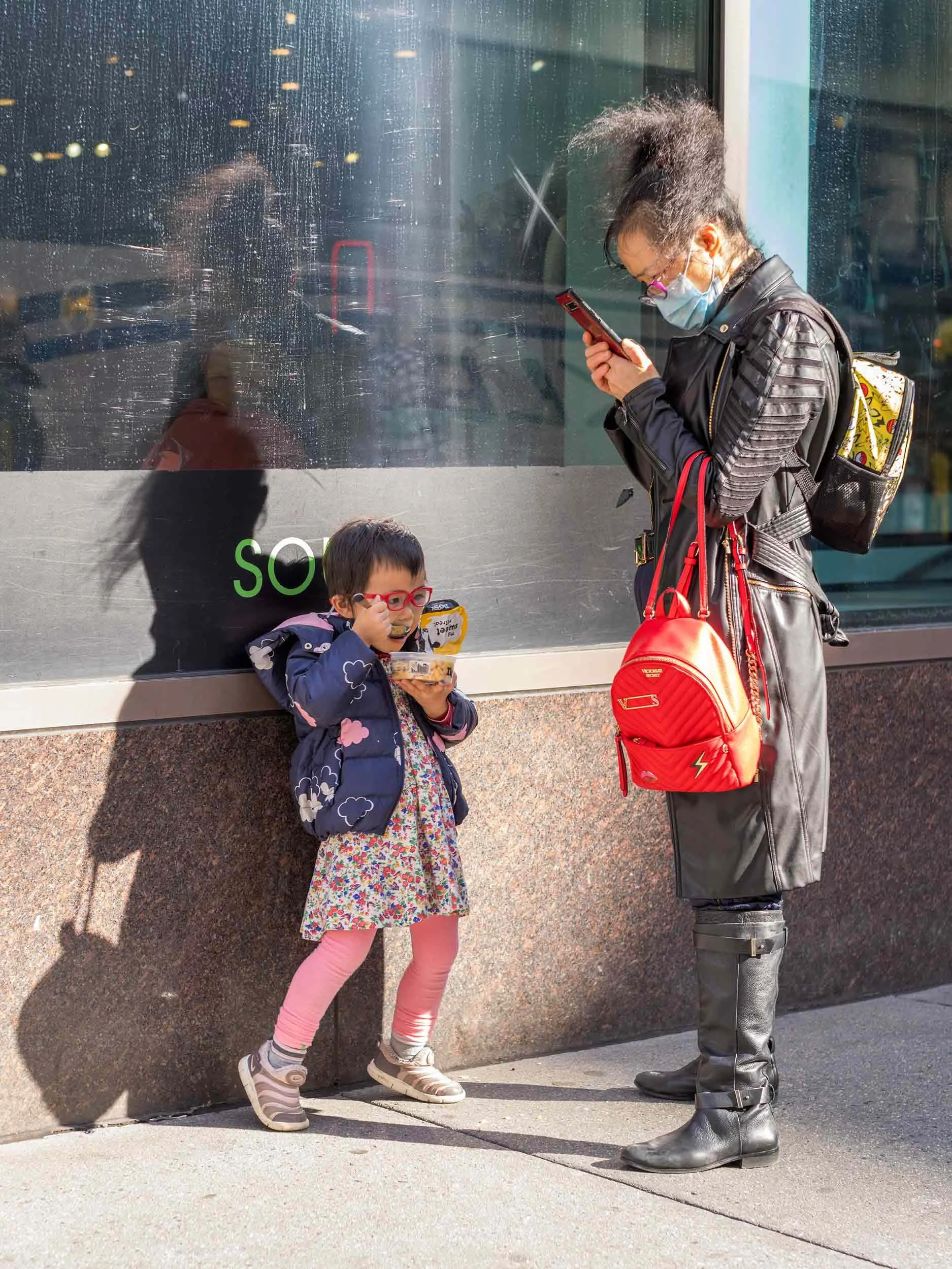 Ein Kind mit roter Brille isst auf dem Gehweg, während eine erwachsene Person neben ihm auf ihr Smartphone schaut.