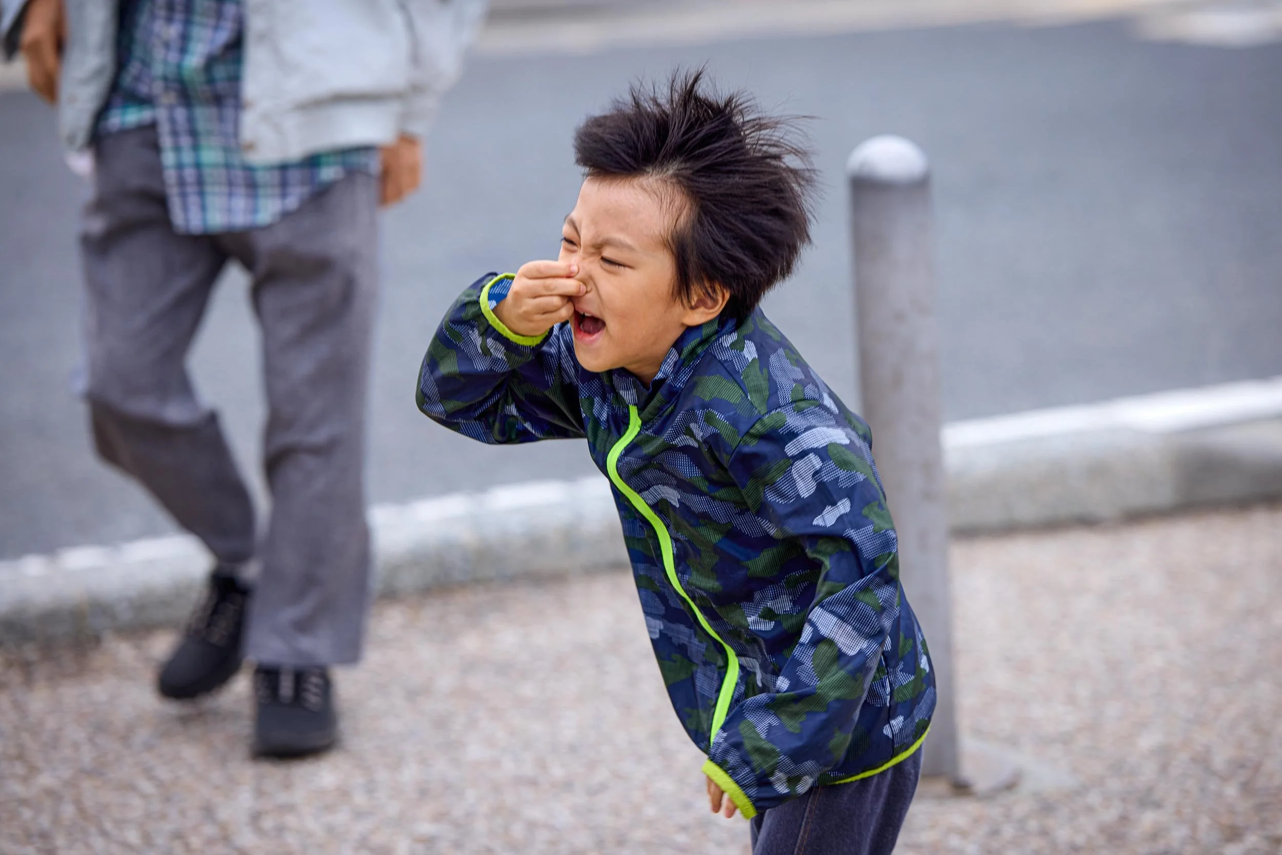 
Kind auf dem Gehweg, hält sich die Nase zu und verzieht dabei das Gesicht zu einer Grimasse.