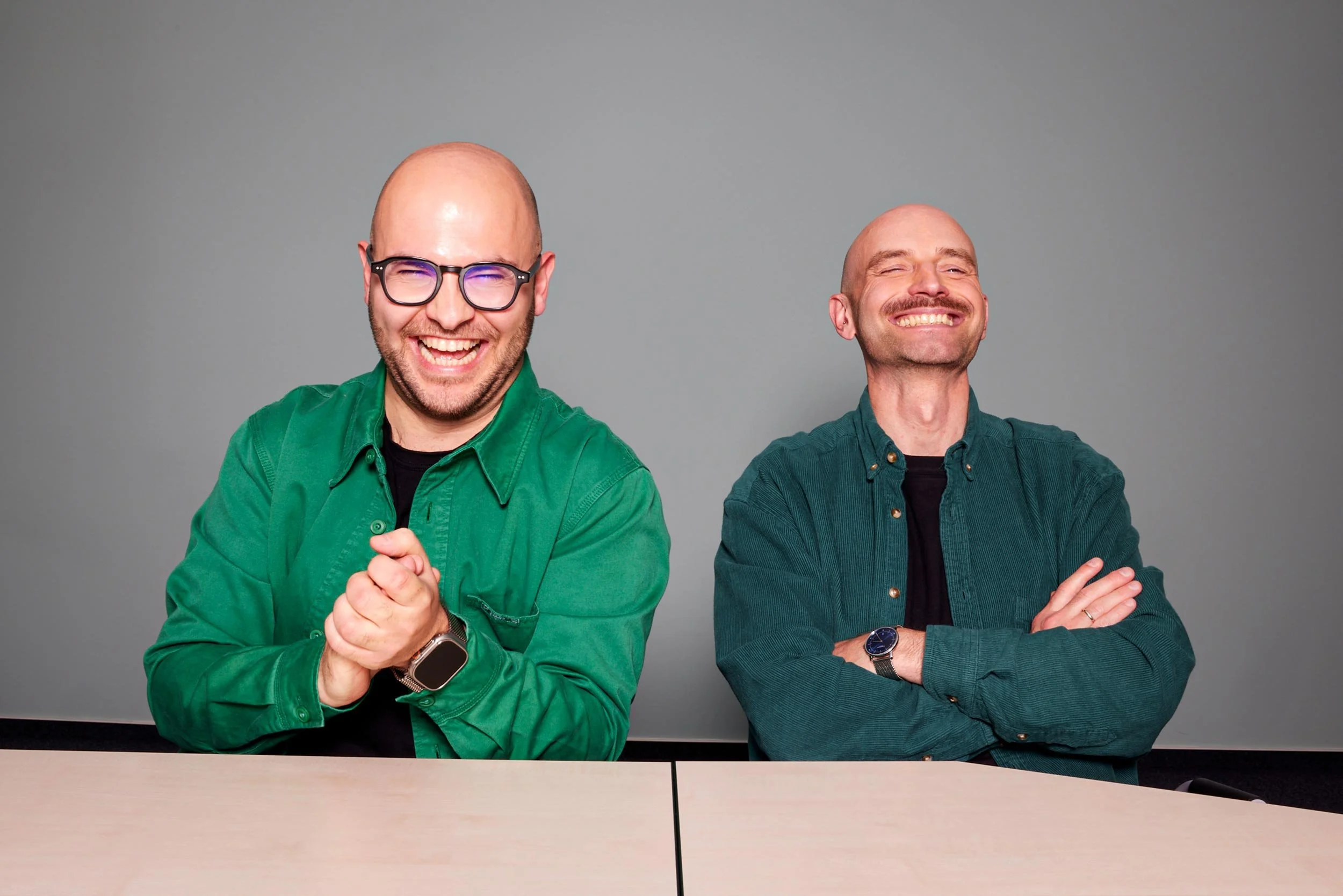 Zwei Männer sitzen an einem Tisch vor grauem Hintergrund und blicken in die Kamera, und lachen herzhaft.
