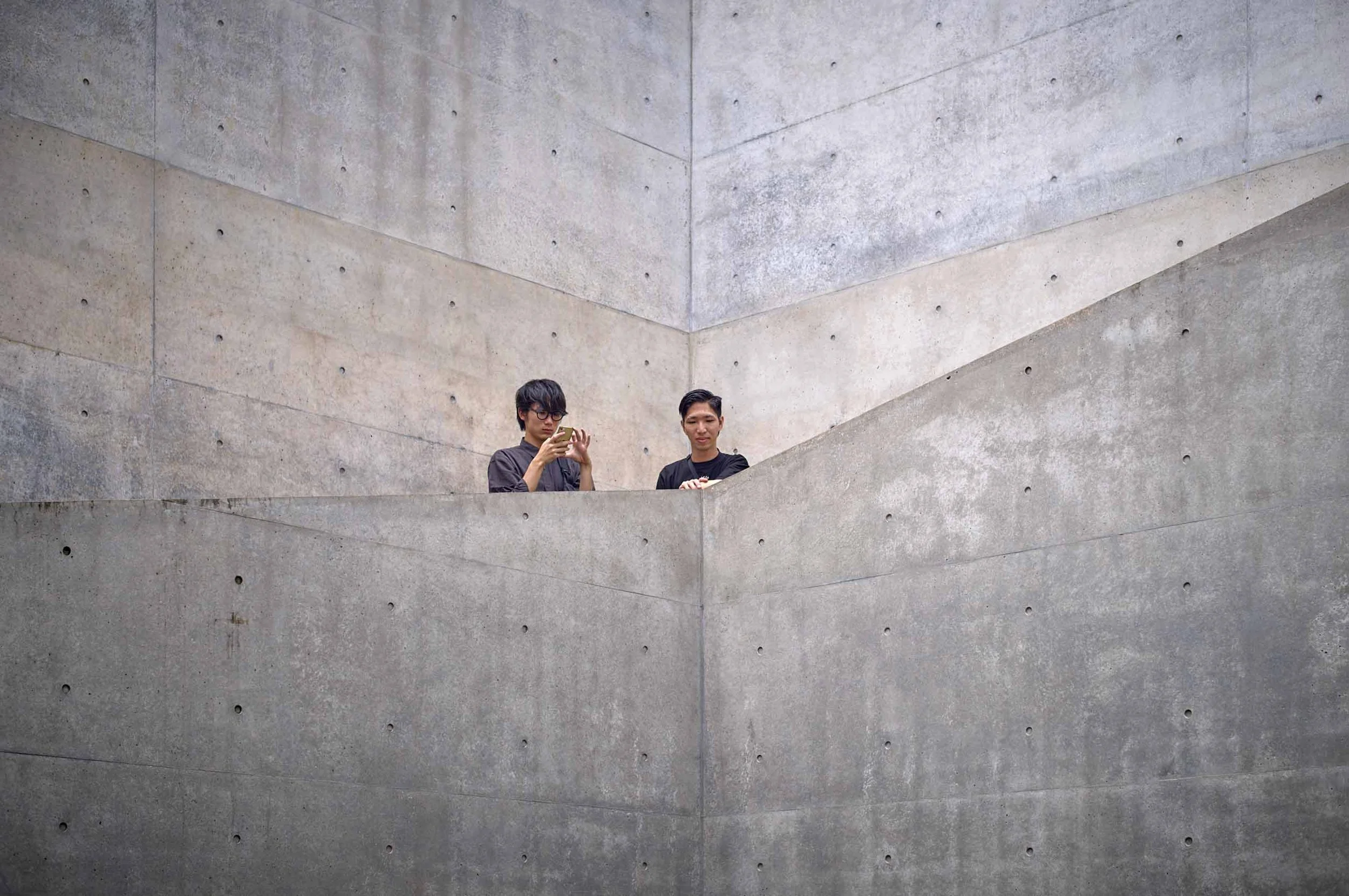 Zwei Männer stehen auf einem Betonbalkon und bedienen Smartphones vor einer Sichtbetonwand von  Tadao Ando,. Chichu Art Museum in naoshima