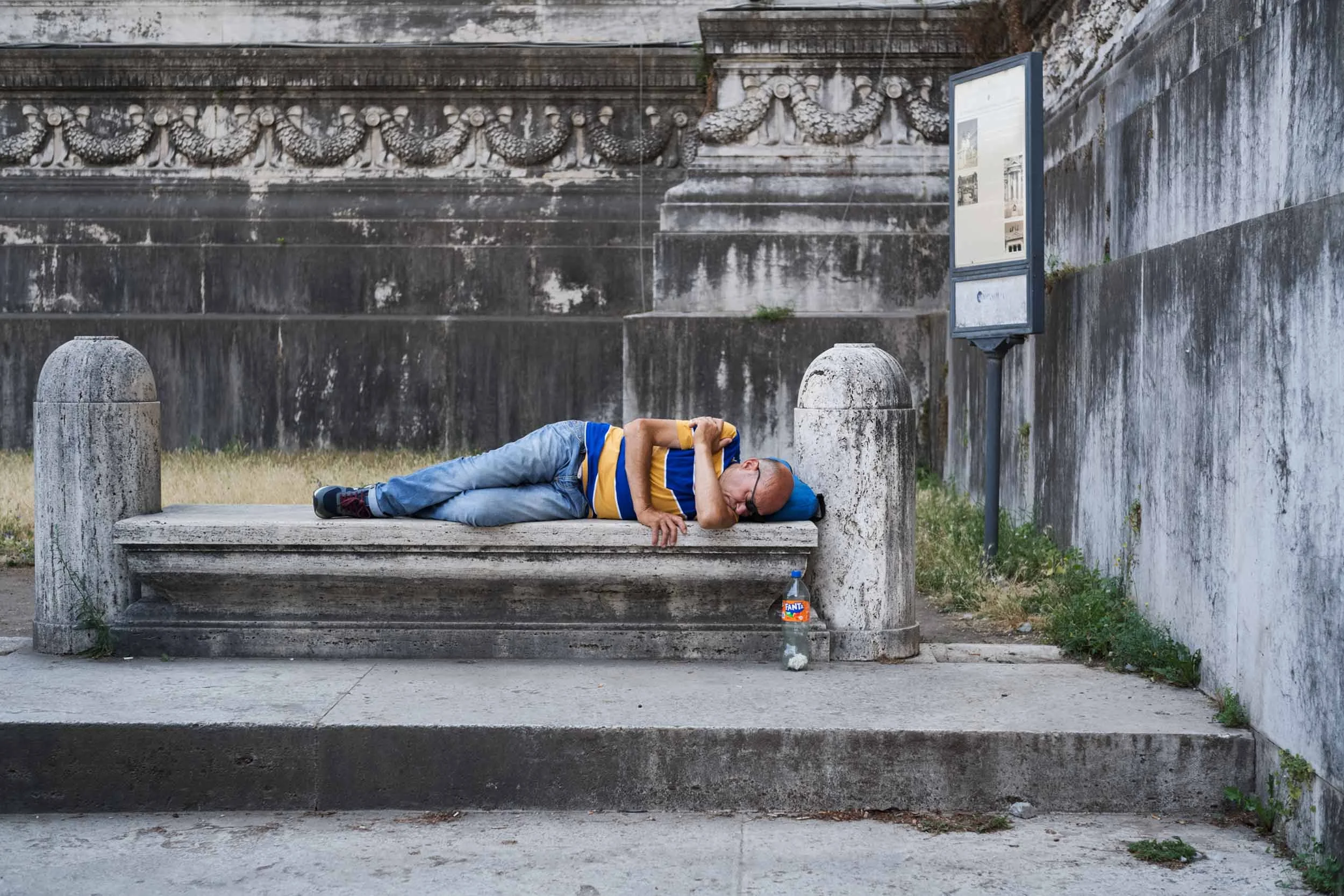 Mann liegt auf einer Steinbank vor einer historischen Steinmauer und schläft, neben der Bank steht eine Plastikflasche.