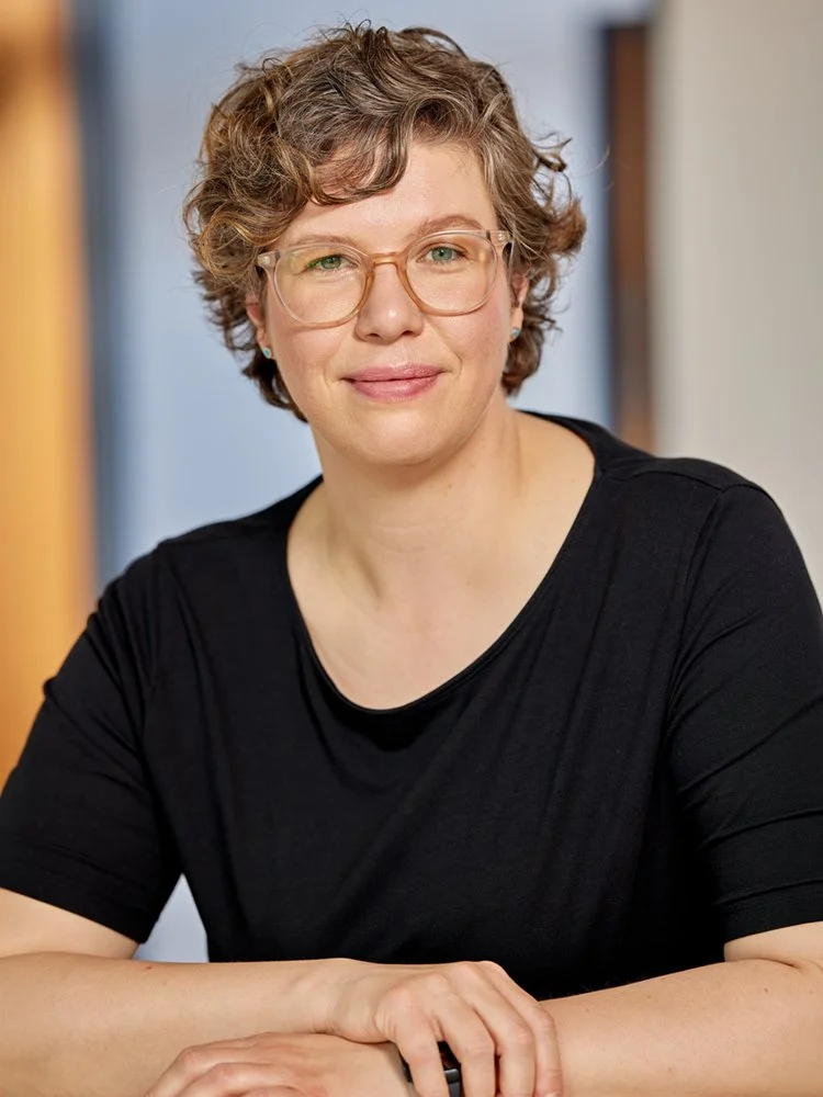 Frau mit Brille und schwarzem Oberteil sitzt an einem Tisch und blickt in die Kamera.