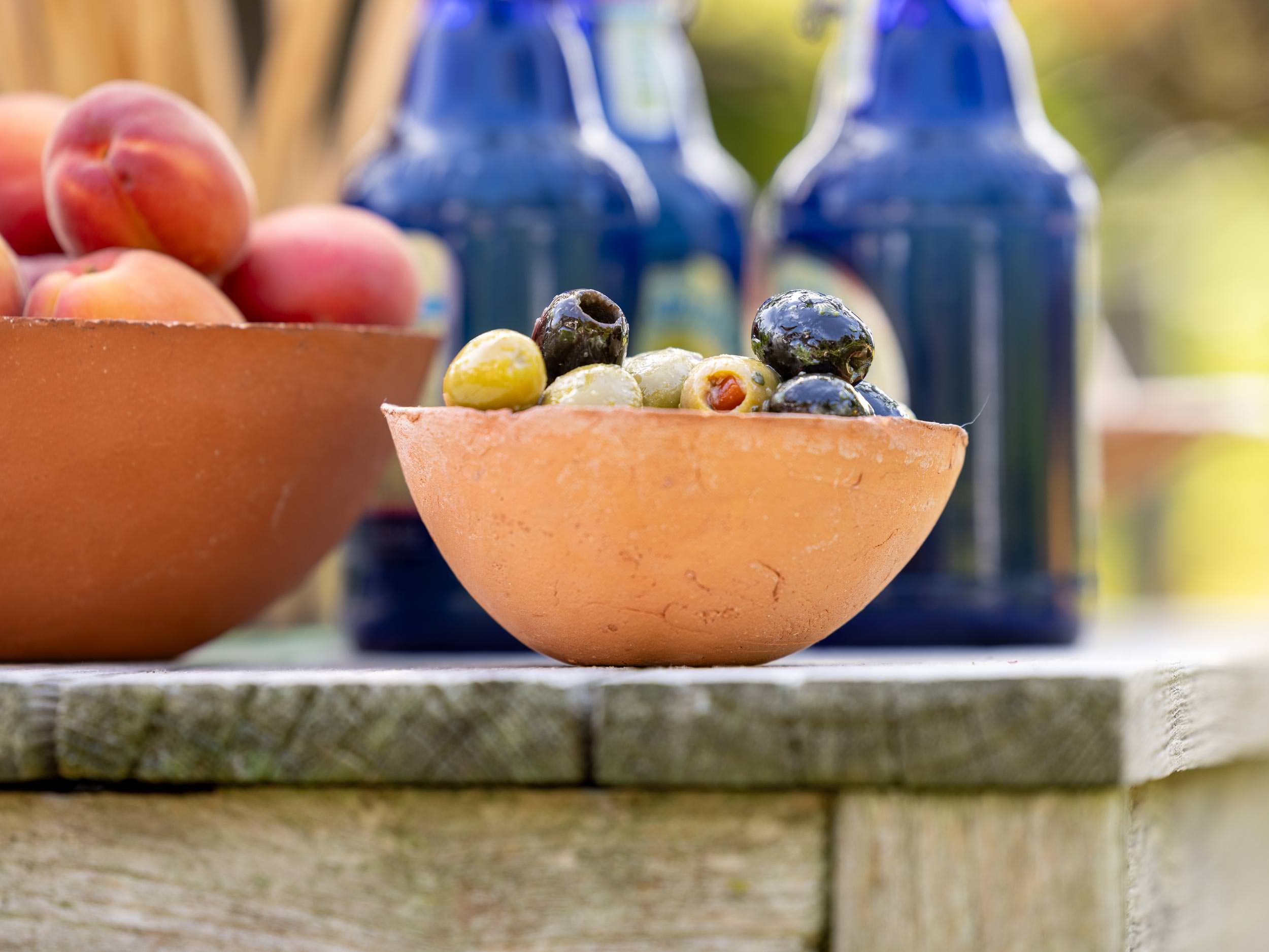 Schale mit grünen und schwarzen Oliven auf Holztisch im Garten, daneben Pfirsiche und blaue Flaschen im Hintergrund.
