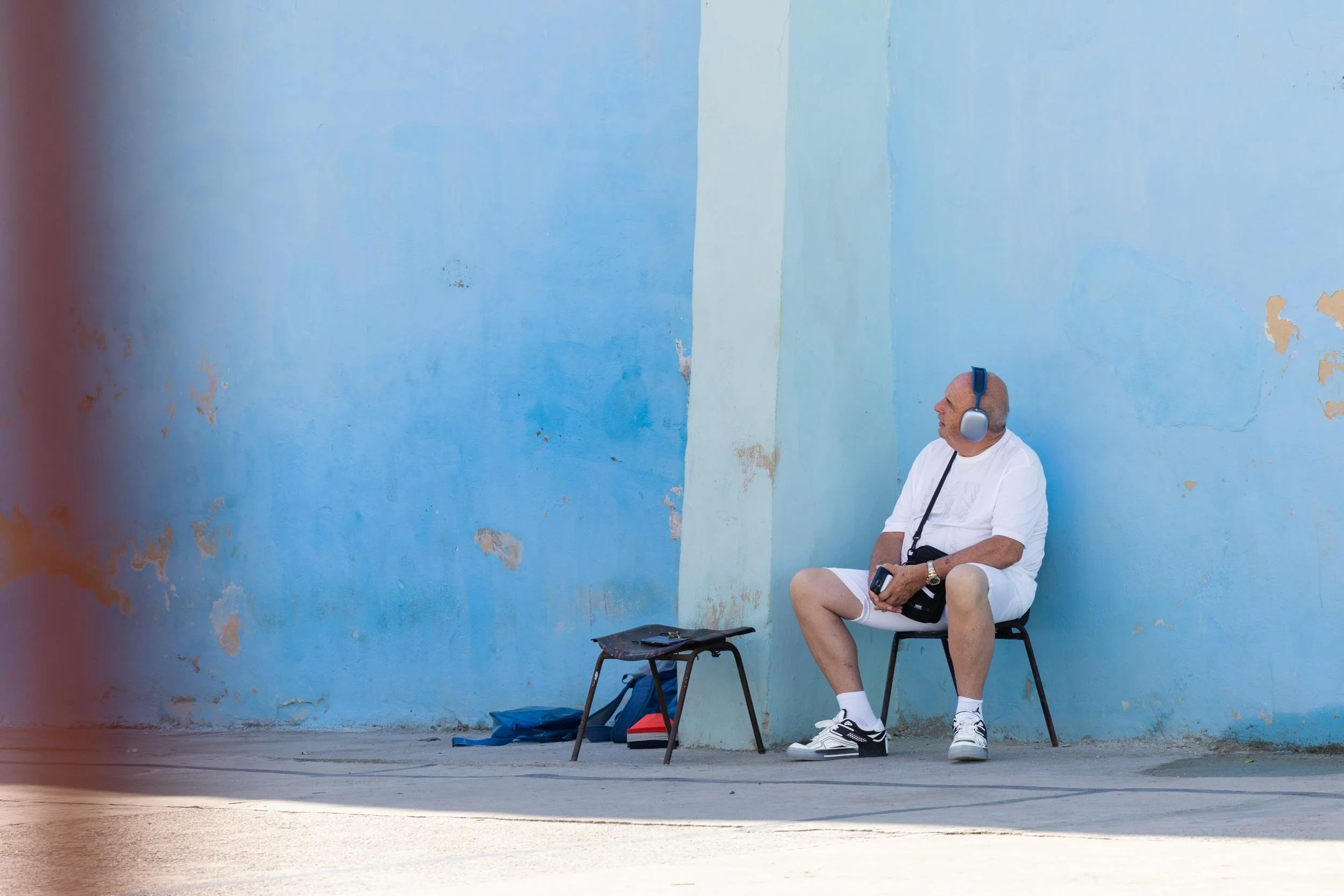 Mann mit Kopfhörern sitzt auf einem Stuhl vor einer blauen Wand, neben ihm steht ein kleiner Tisch.