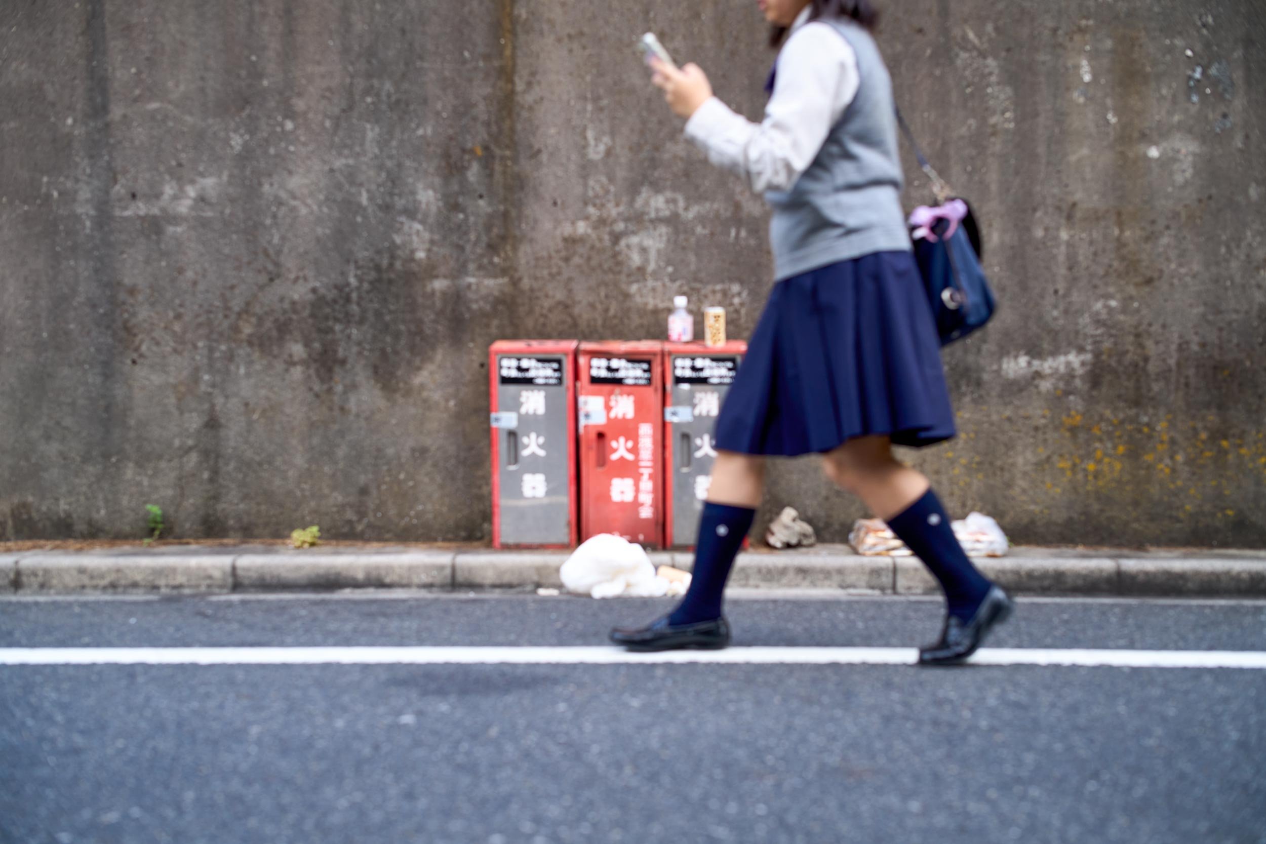 Schülerin in Uniform läuft an einer Betonwand entlang und schaut auf ihr Smartphone.