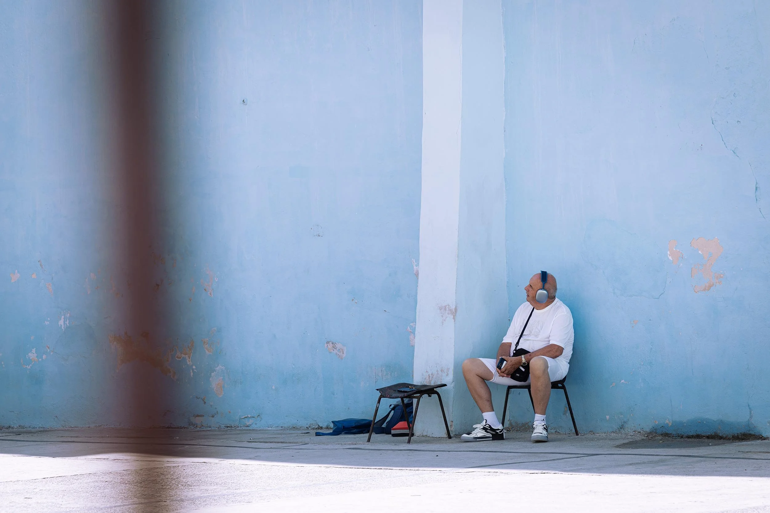 Ein Mann mit Kopfhörern sitzt allein auf einem Stuhl vor einer großflächigen hellblauen Wand an einer Straße in Kuba, 2022.