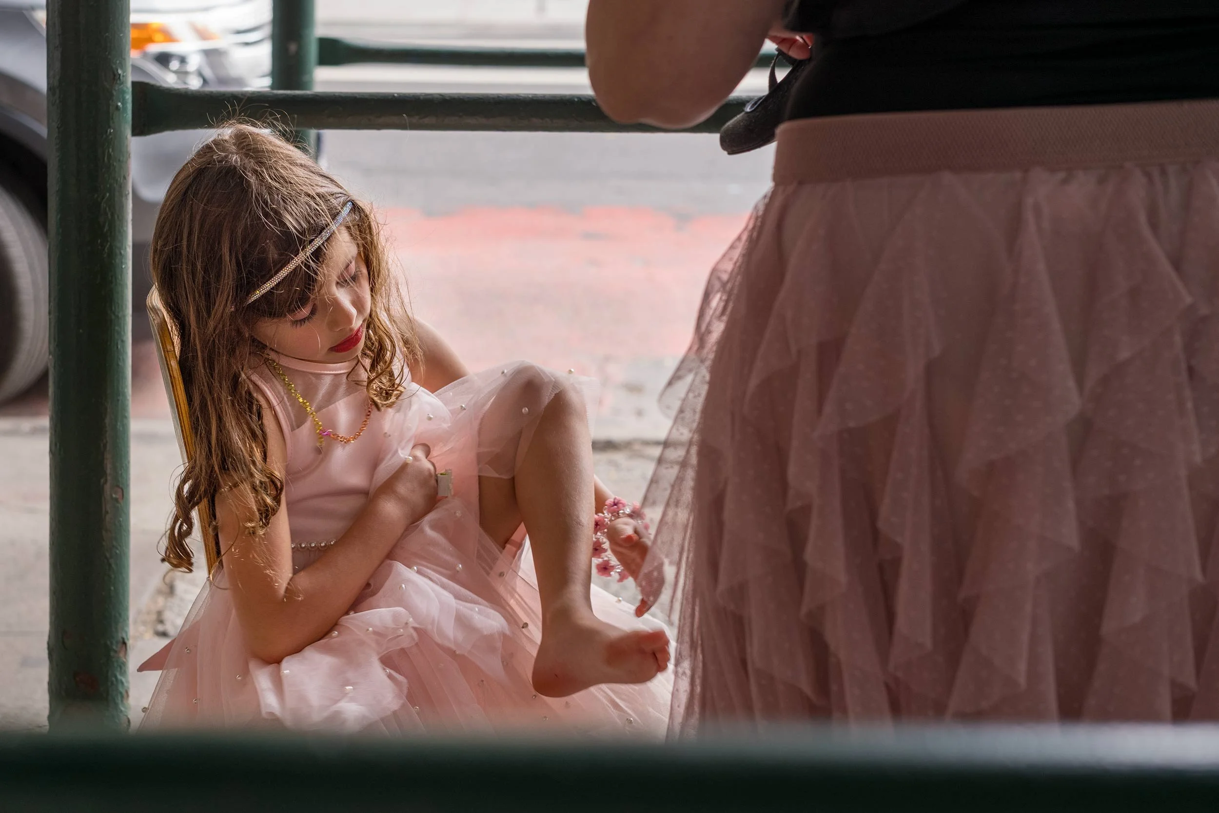 Ein kleines Mädchen im rosa Kleid sitzt auf dem Gehweg und betrachtet seinen Fuß, während eine erwachsene Person neben ihm steht.