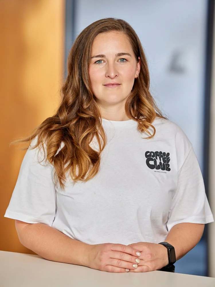 Frau mit langen Haaren und weißem T-Shirt steht an einem Tisch und blickt in die Kamera.