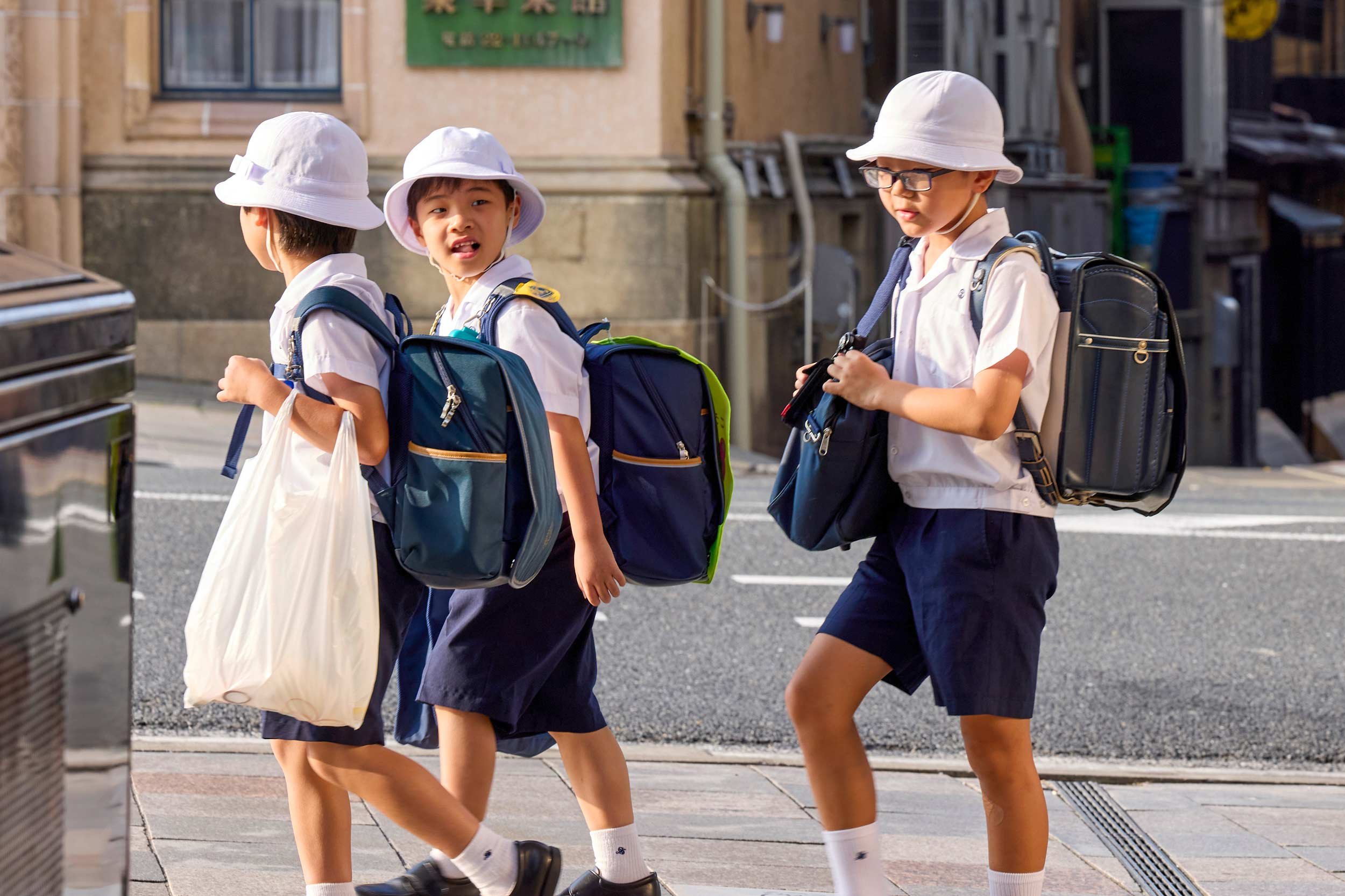 Drei Schulkinder mit Rucksäcken und Hüten gehen über eine Straße in Kyoto, ein Kind trägt eine Tasche.