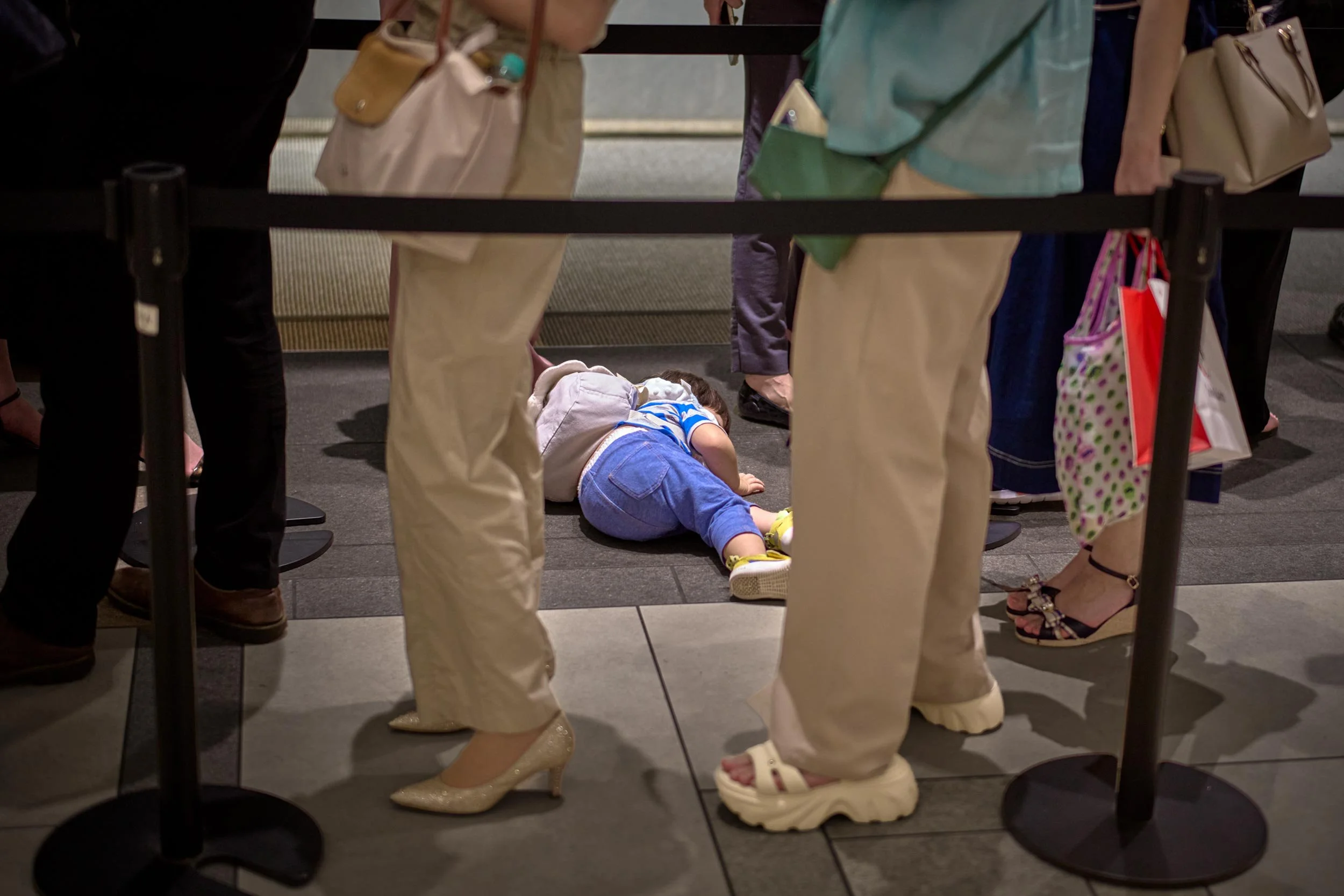 Kind liegt am Boden zwischen Absperrbändern, umgeben von stehenden Personen in einer Warteschlange