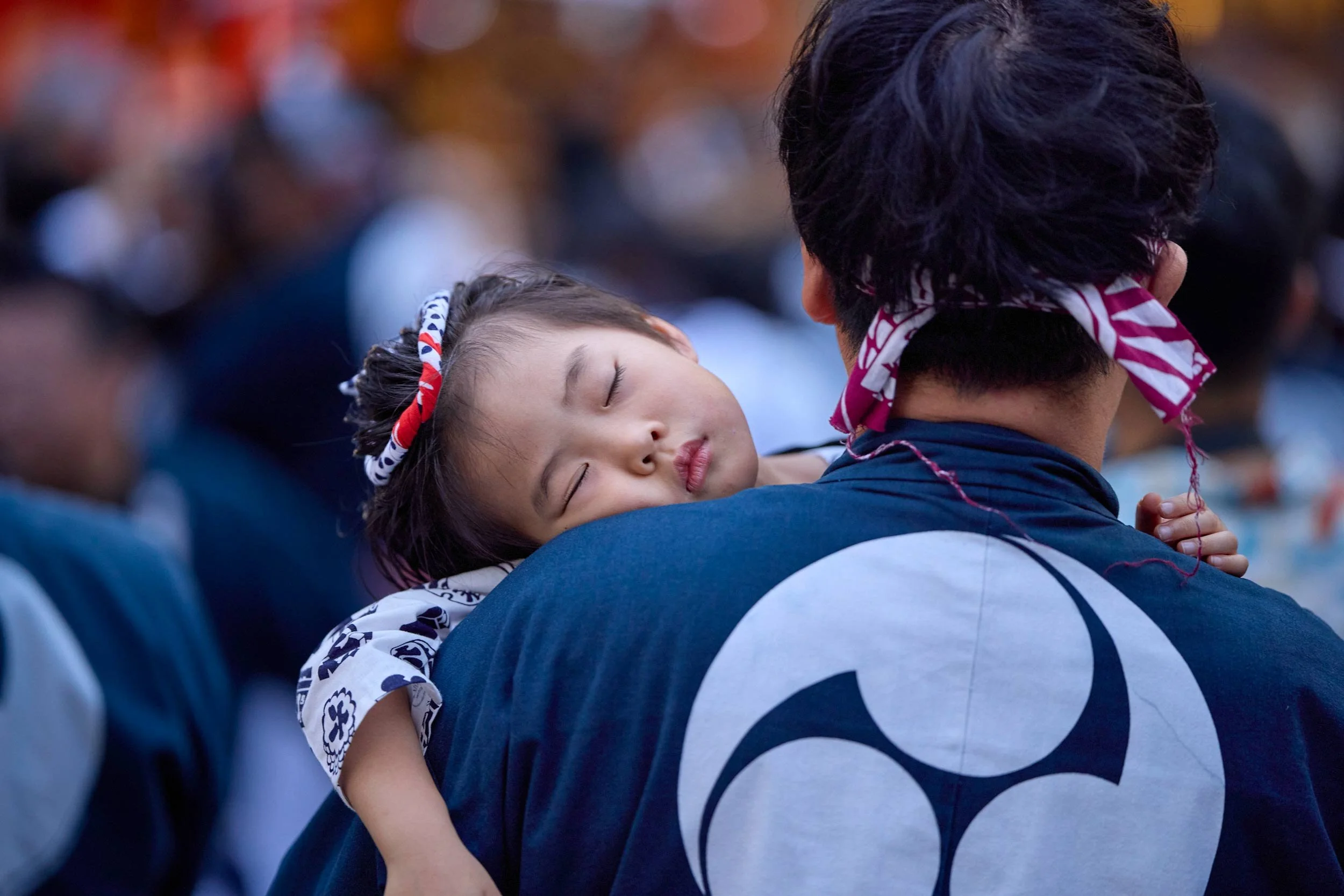 Kind schläft mit geschlossenen Augen auf der Schulter eines Erwachsenen, beide tragen traditionelle Kleidung auf einem Straßenfest in Tokio.
