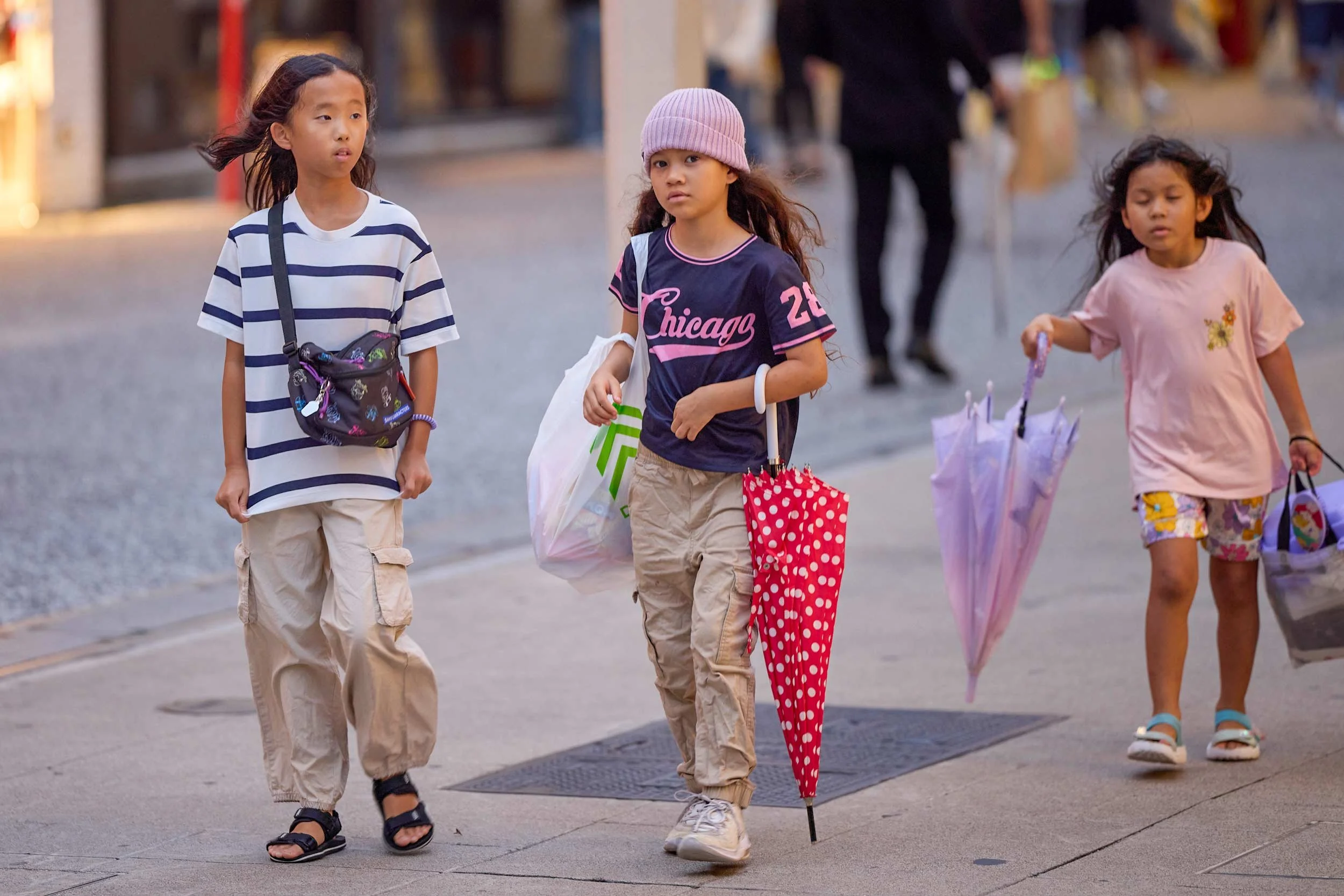 Drei Kinder gehen durch eine Fußgängerzone in Hiroshima, sie tragen Einkaufstaschen und einen Regenschirm.