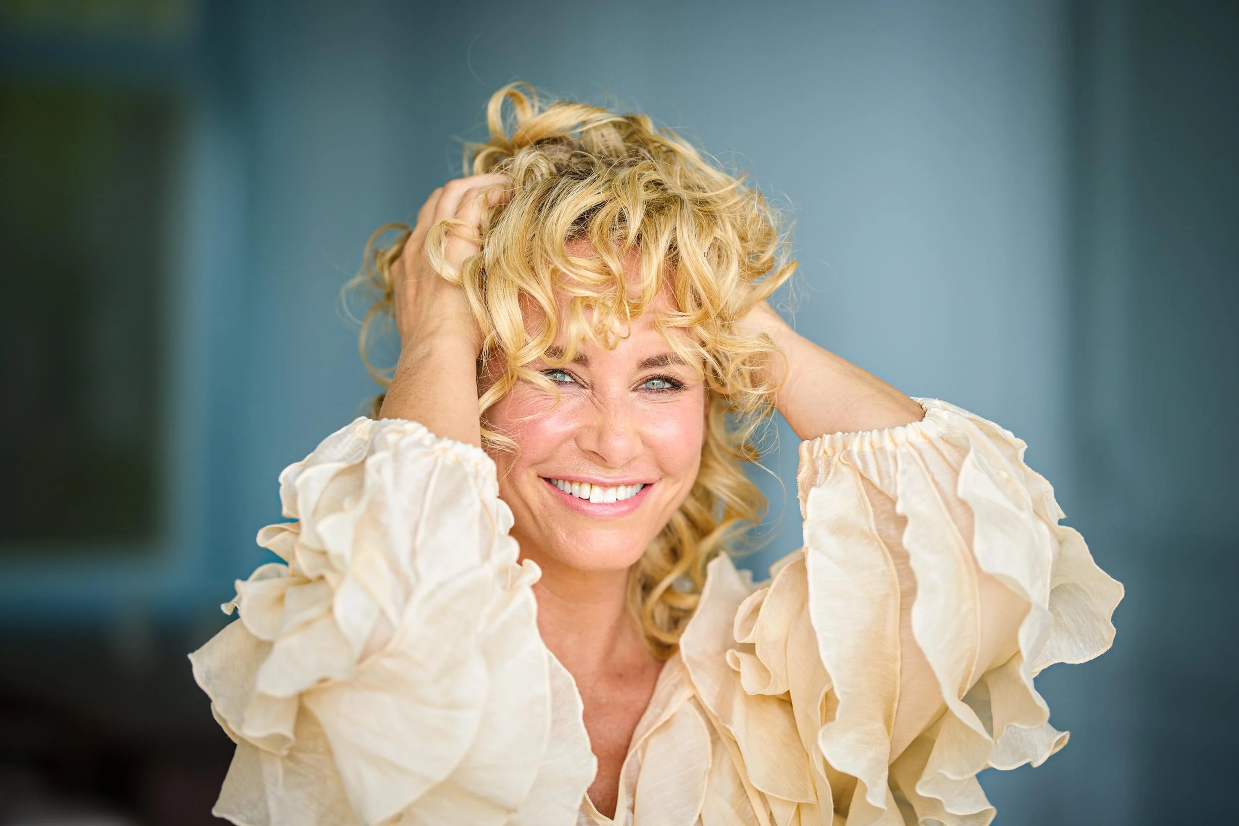 Blonde Frau mit lockigem Haar lächelt und hält beide Hände in ihren Locken.