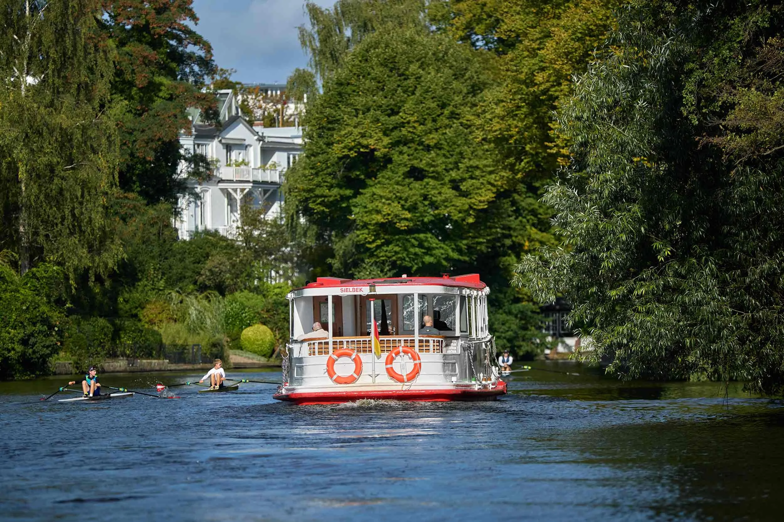 Alsterschiff Sielbek auf einem grünen Hamburger Alsterkanal, begleitet von Ruderern und historischen Villen am Ufer, aus der Fotoserie „Alster Paradise“ von David Goltz.