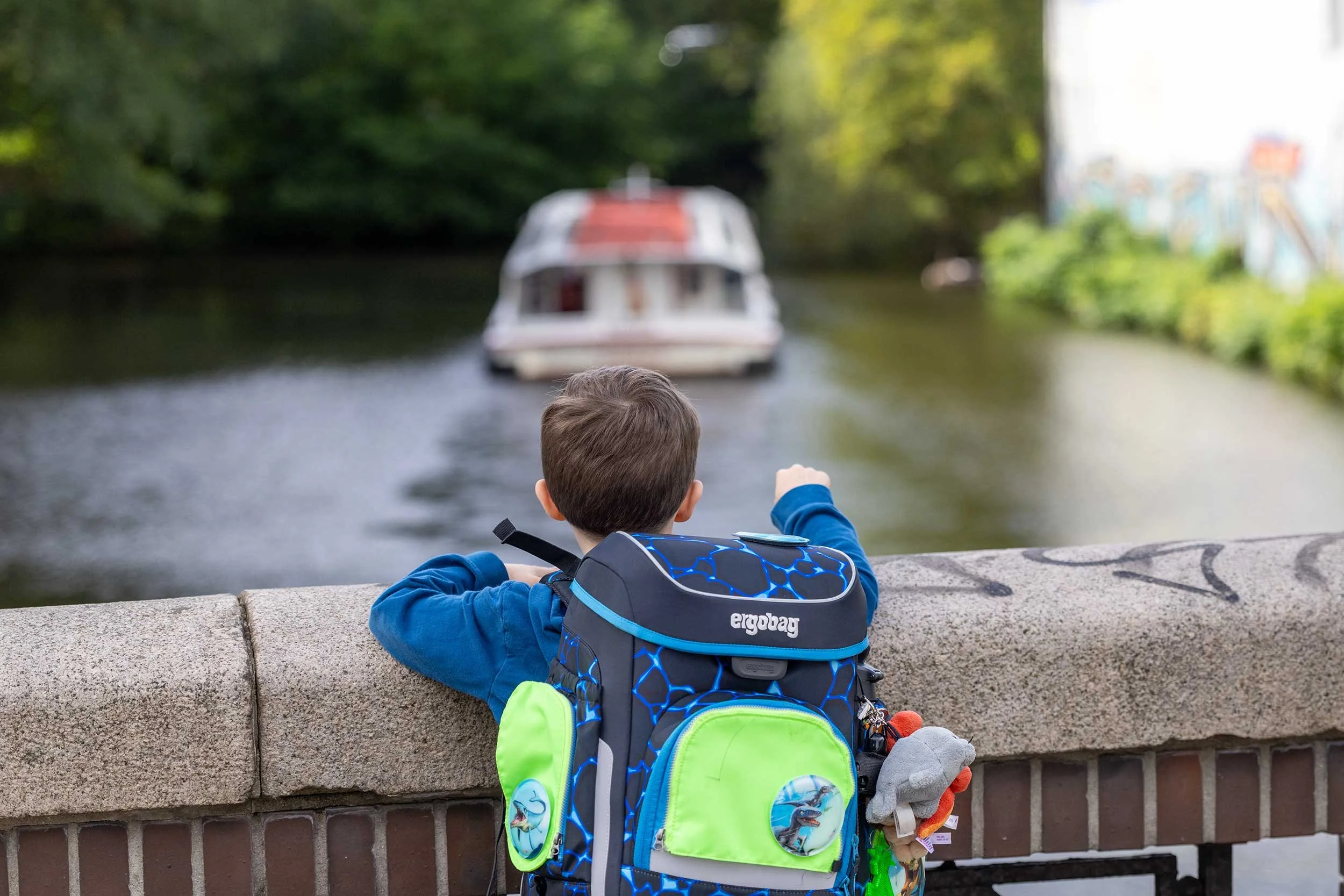 Ein Kind mit auffällig blau-grünem Schulranzen lehnt an einer Mauer und schaut einem vorbeifahrenden Alsterschiff hinterher. Die geringe Schärfentiefe lenkt den Blick auf das Kind, während Schiff, Wasser und grüne Uferlandschaft weich im Hintergrund 