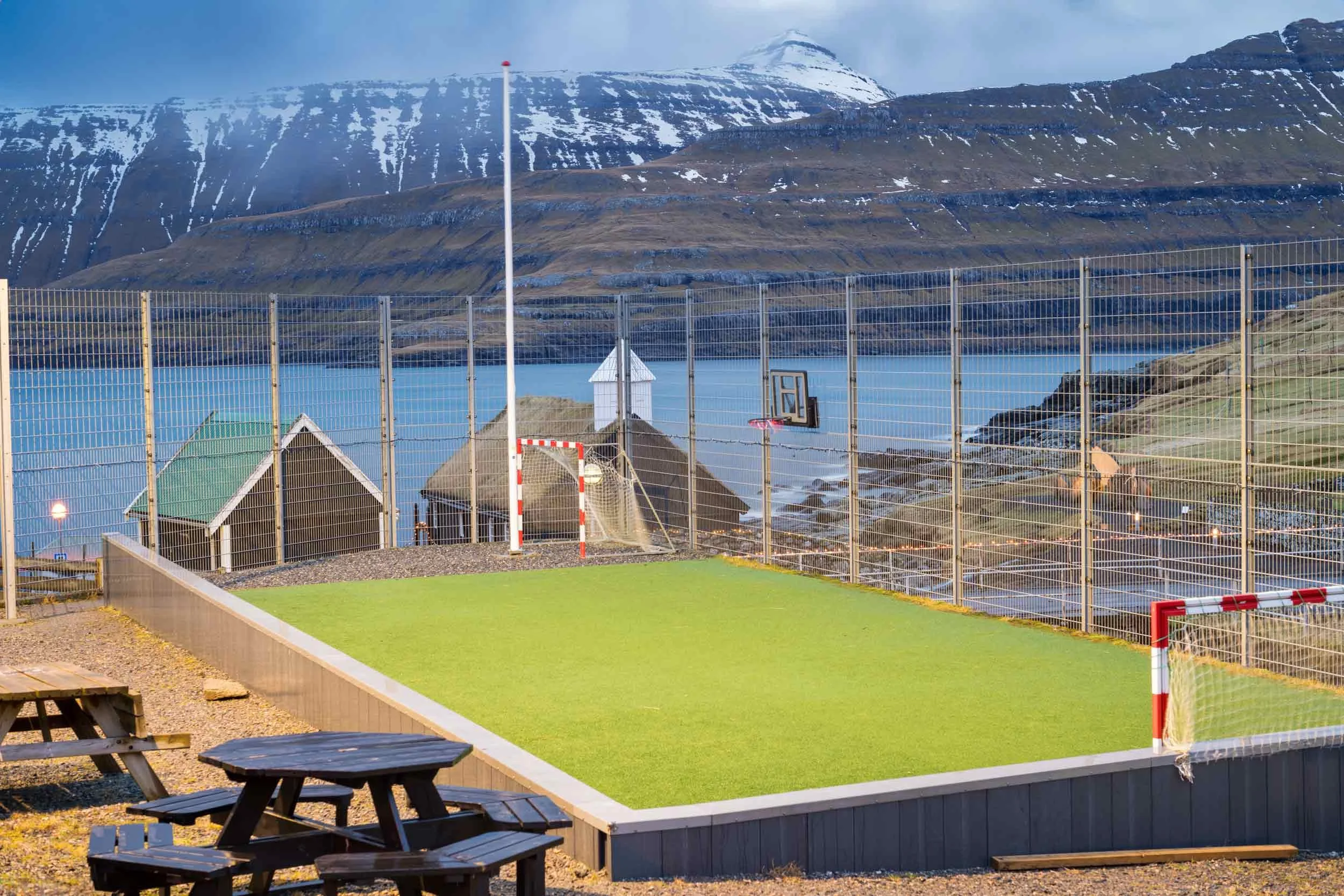 Das Bild zeigt einen kleinen Fußballplatz auf den Färöer-Inseln, umgeben von einer malerischen, schroffen Landschaft mit Bergen und Seen.
