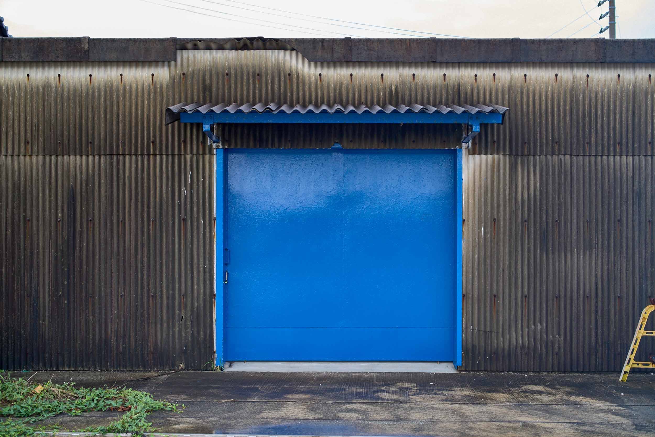 Blaues Metalltor in einer grauen Wellblechwand mit kleinem Dach darüber.