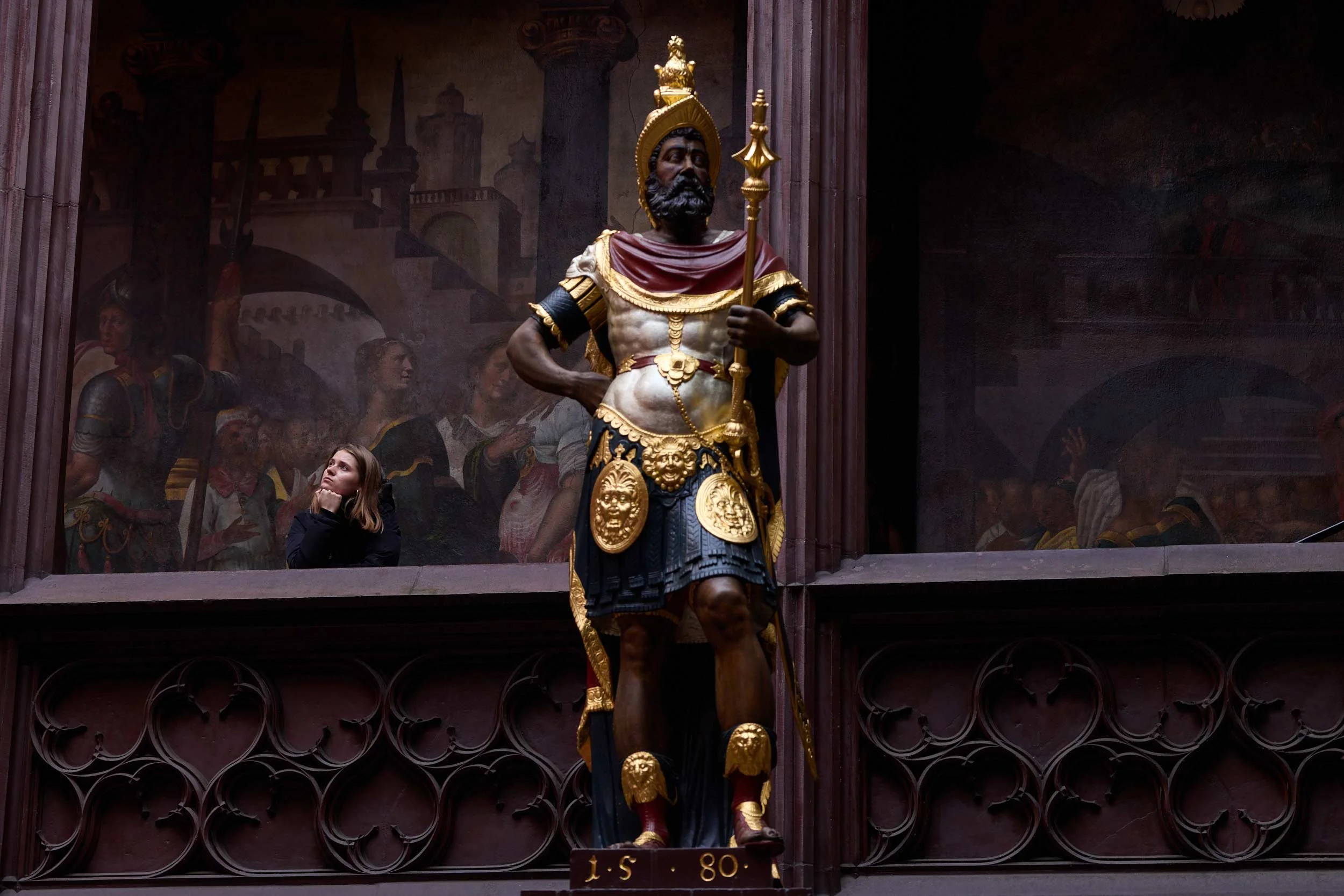 Grosse bemalte Statue steht in einem Innenraum, dahinter eine Person an einer Brüstung.