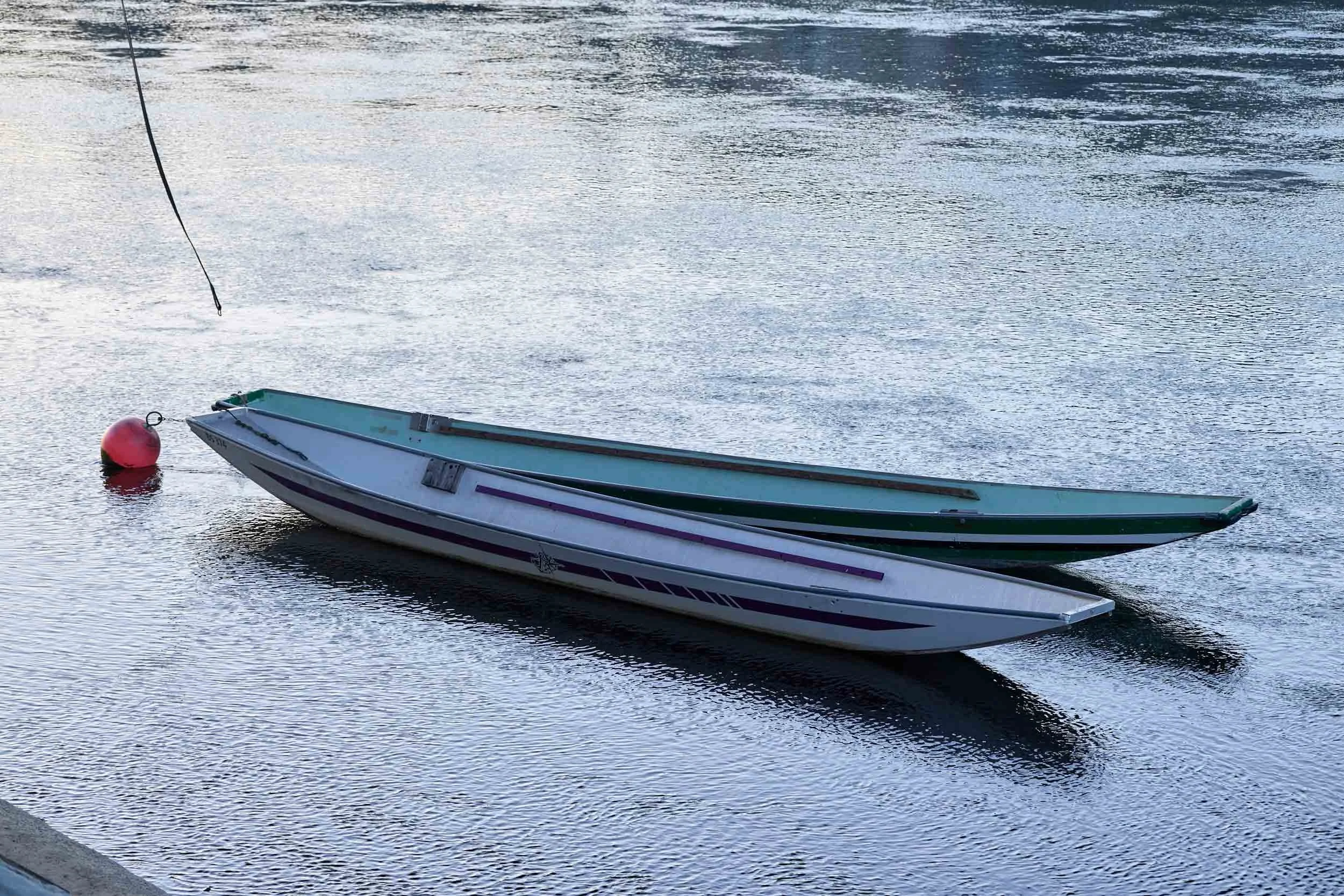 Zwei kleine Boote liegen nebeneinander im Wasser und sind an einer roten Boje befestigt.
