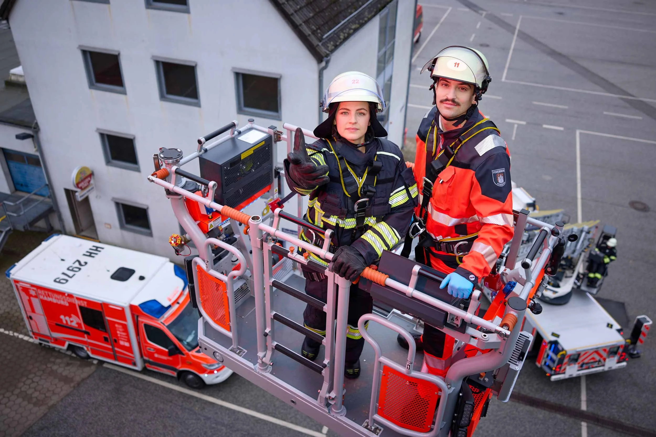 Feuerwehrkräfte auf einer Drehleiter über einem Feuerwehrhof mit Einsatzfahrzeugen