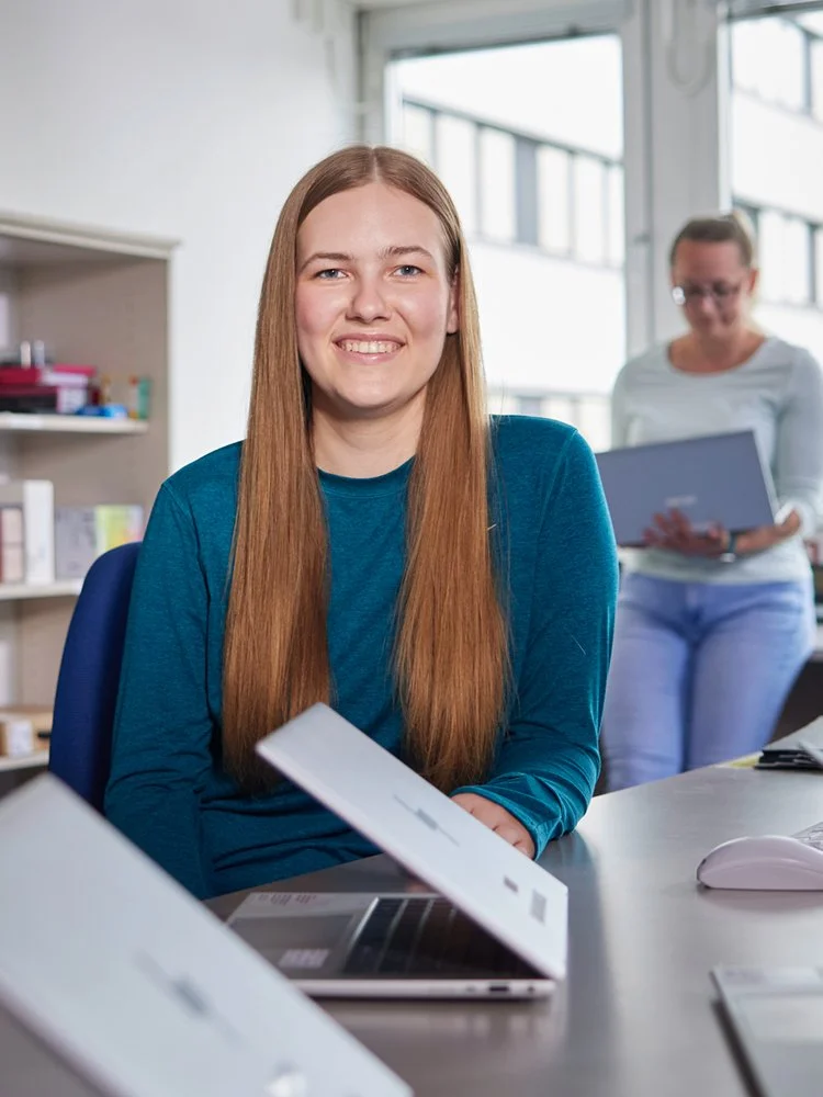 Frau sitzt an einem Schreibtisch in einem Büro, während im Hintergrund eine weitere Person mit einem Laptop steht.