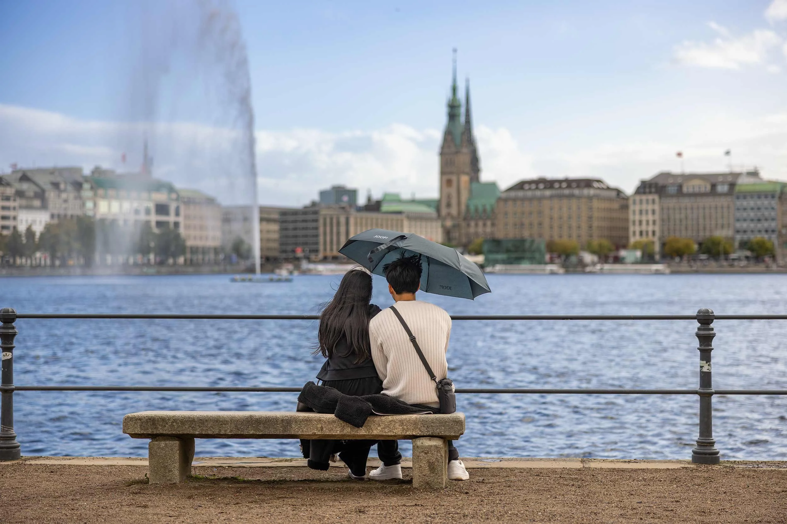 Ein Paar sitzt unter einem Regenschirm auf einer Bank an der Hamburger Binnenalster und blickt über das Wasser auf die Innenstadt, aus der Fotoserie „Alster Paradise“ von David Goltz.