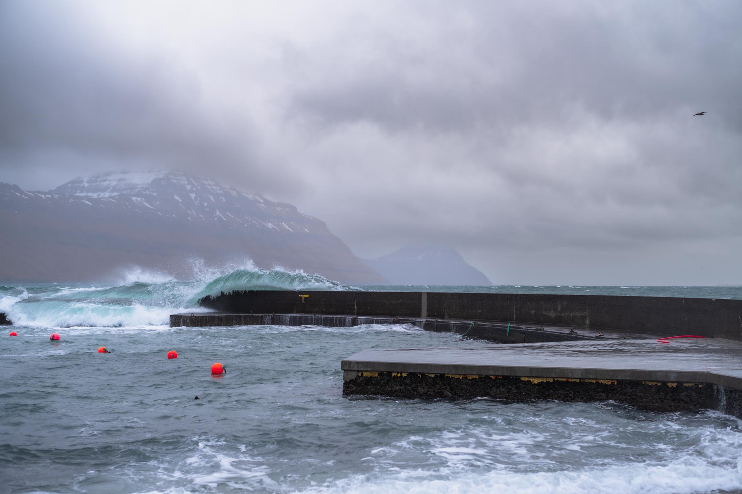 Welle bricht gegen eine hafenmauer vor berglandschaft unter bewolktem himmel, faeroer
