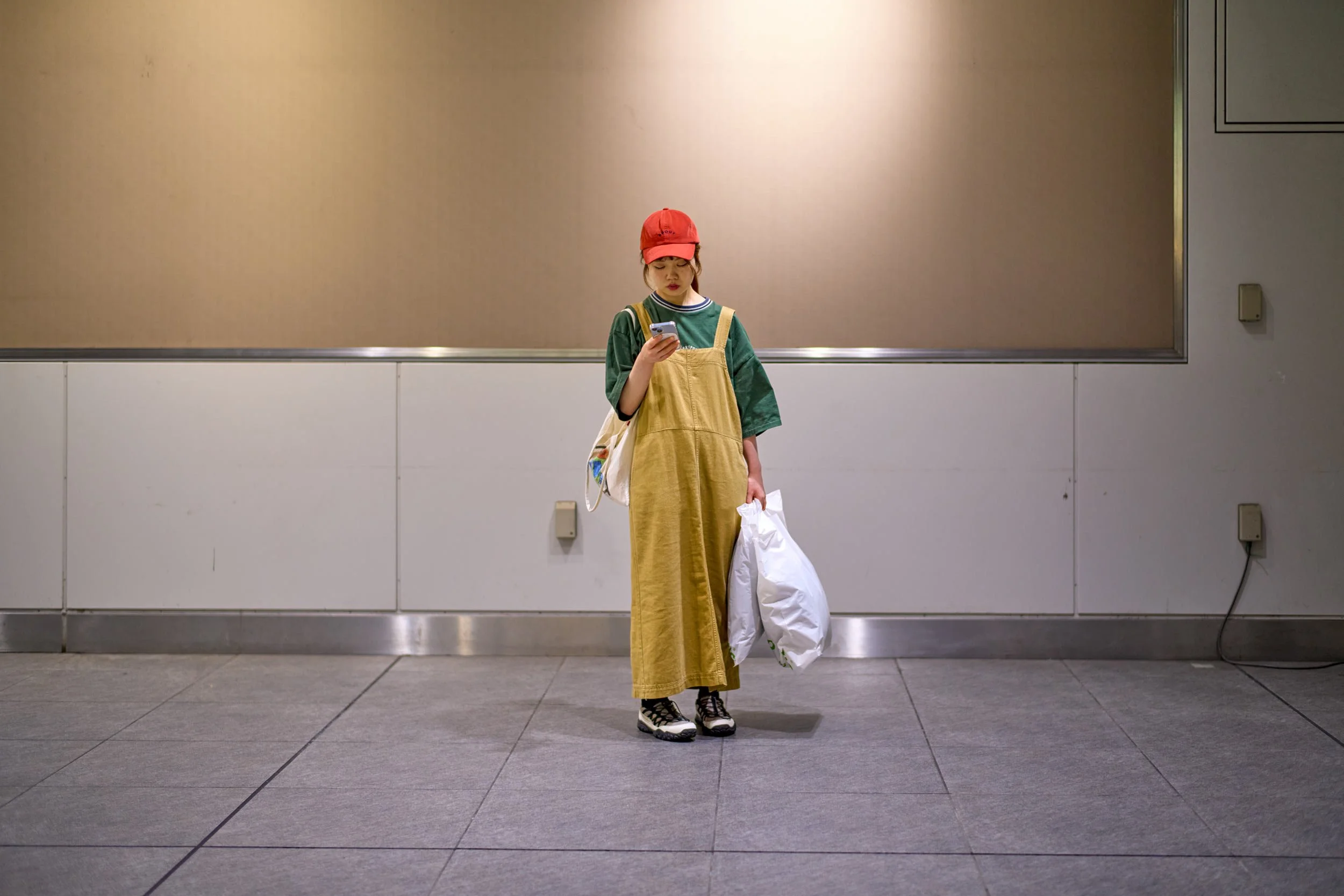 Frau mit roter Kappe steht in einer Unterführung, schaut auf ihr Smartphone und hält Einkaufstüten