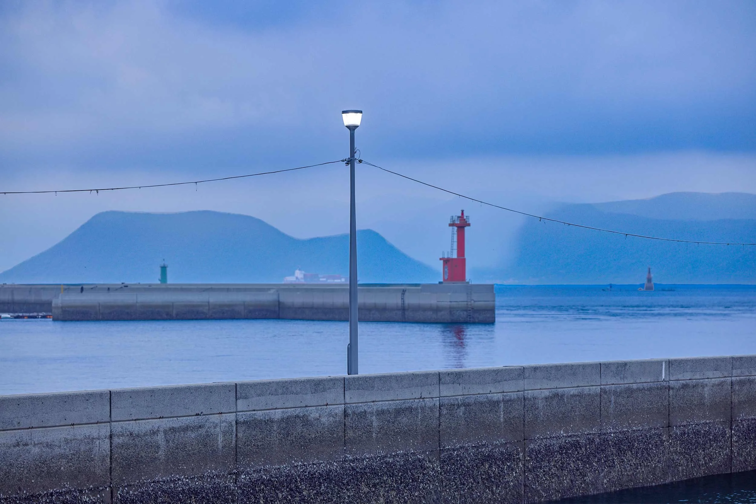 Hafenmauer mit Laterne im Vordergrund und rotem Leuchtturm im Hintergrund auf Naoshima.