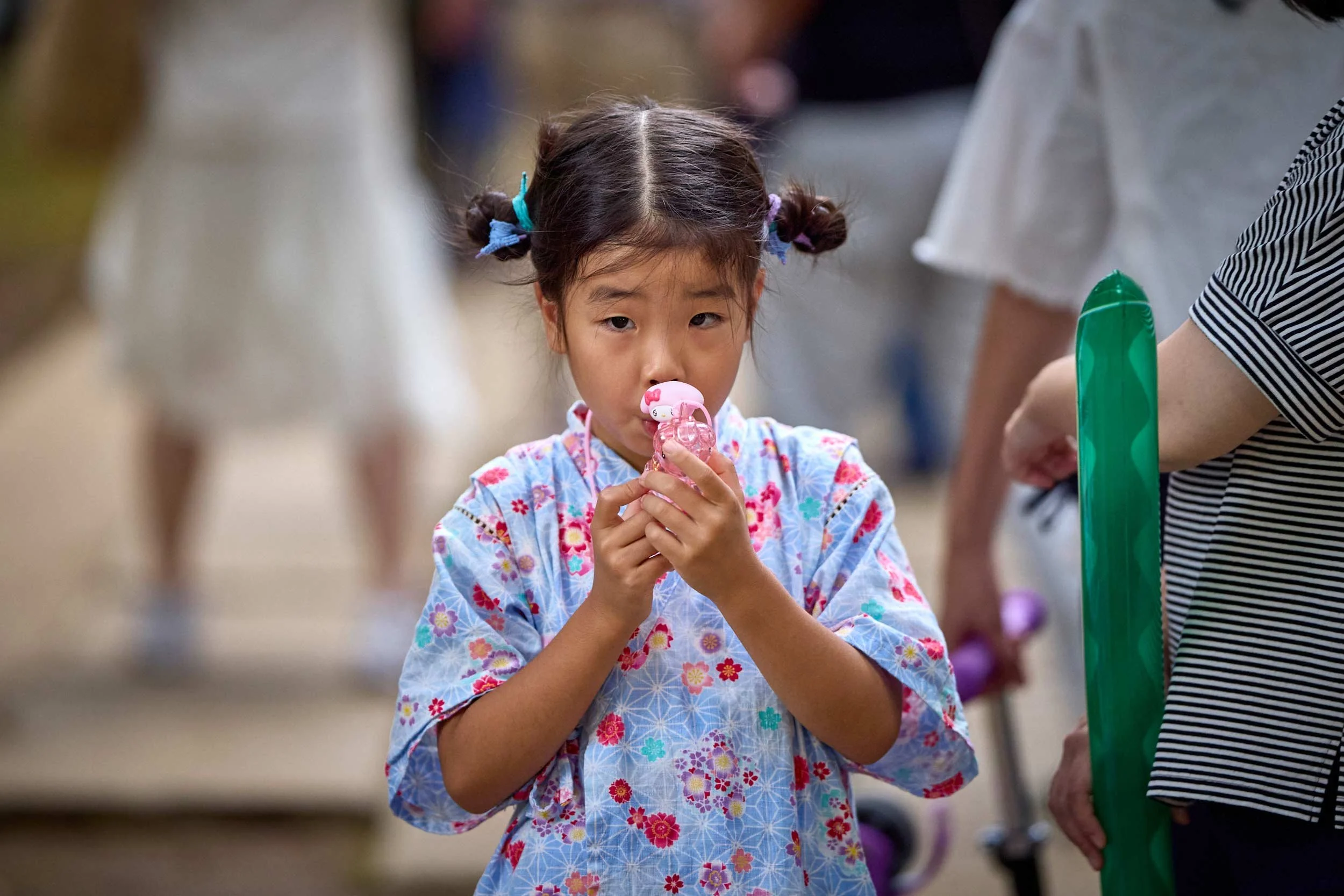 Kind in einem gemusterten Yukata hält ein kleines Spielzeug und trinkt daraus, Erwachsene und weitere Personen stehen im Hintergrund.