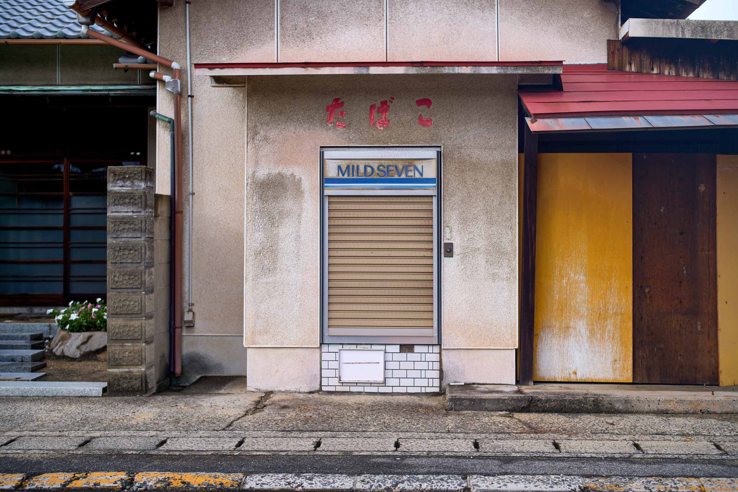 Geschlossenes Verkaufsfenster mit Rolladen und dem Schriftzug ‚MILD SEVEN‘ an einer japanischen Hausfassade, daneben eine gelbe Tür.