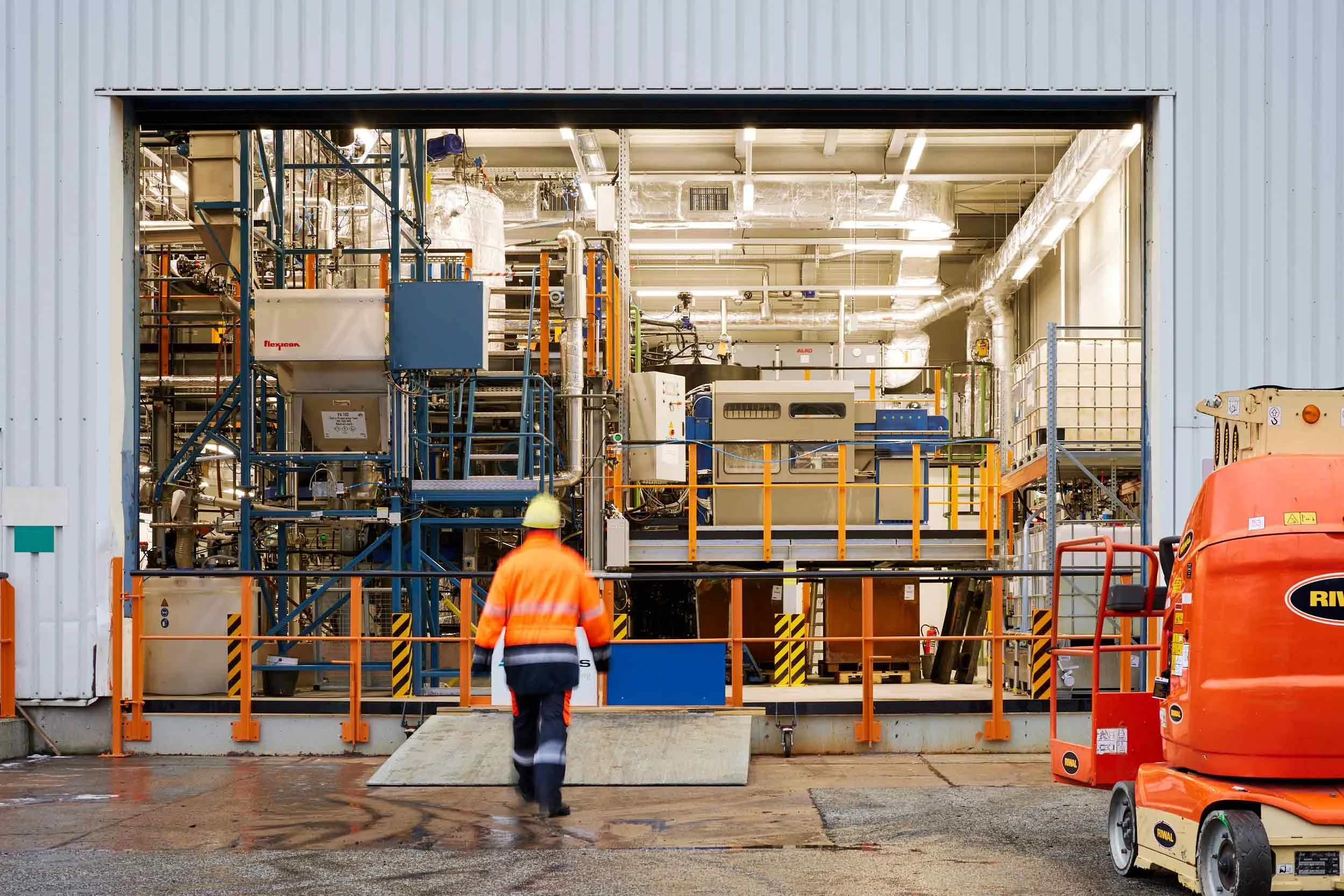 Blick in die Produktionshalle durch das geöffnete Rolltor – Industriearchitektur, Technikvielfalt und menschliche Präsenz im Einklang.