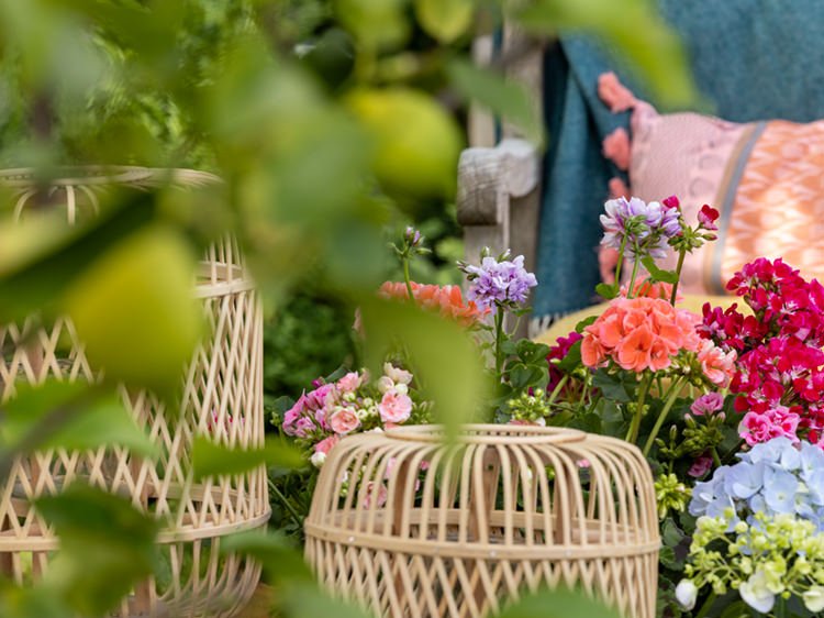 Blumenarrangement und Rattan-Dekoration in Gartenlounge mit Kissen und Pflanzen.