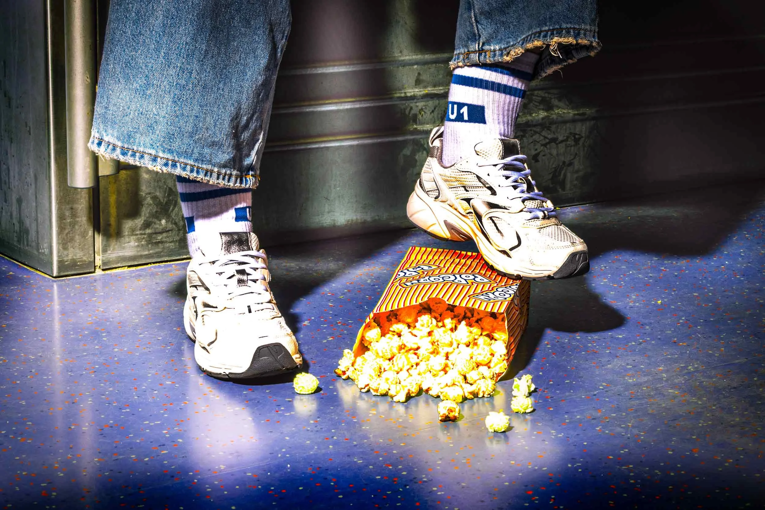 Detailaufnahme von weißen Sneakern und Hochbahn-U1-Socken im U-Bahn-Wagen mit verschüttetem Popcorn.