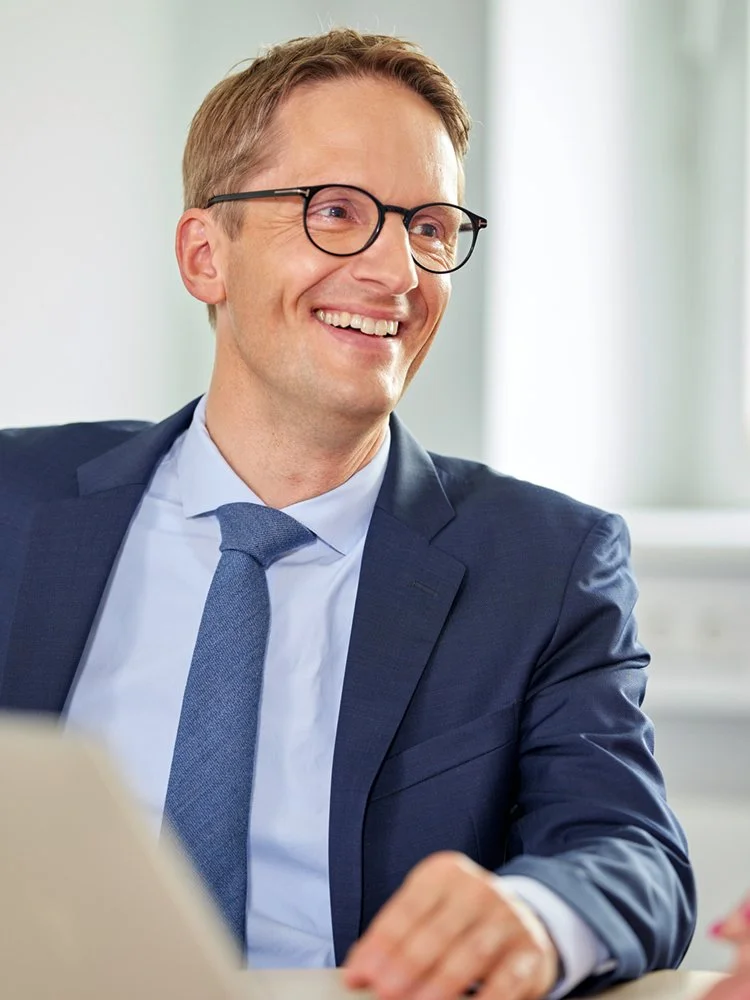 Mann mit Brille, Anzug und Krawatte sitzt an einem Tisch und blickt zur Seite.