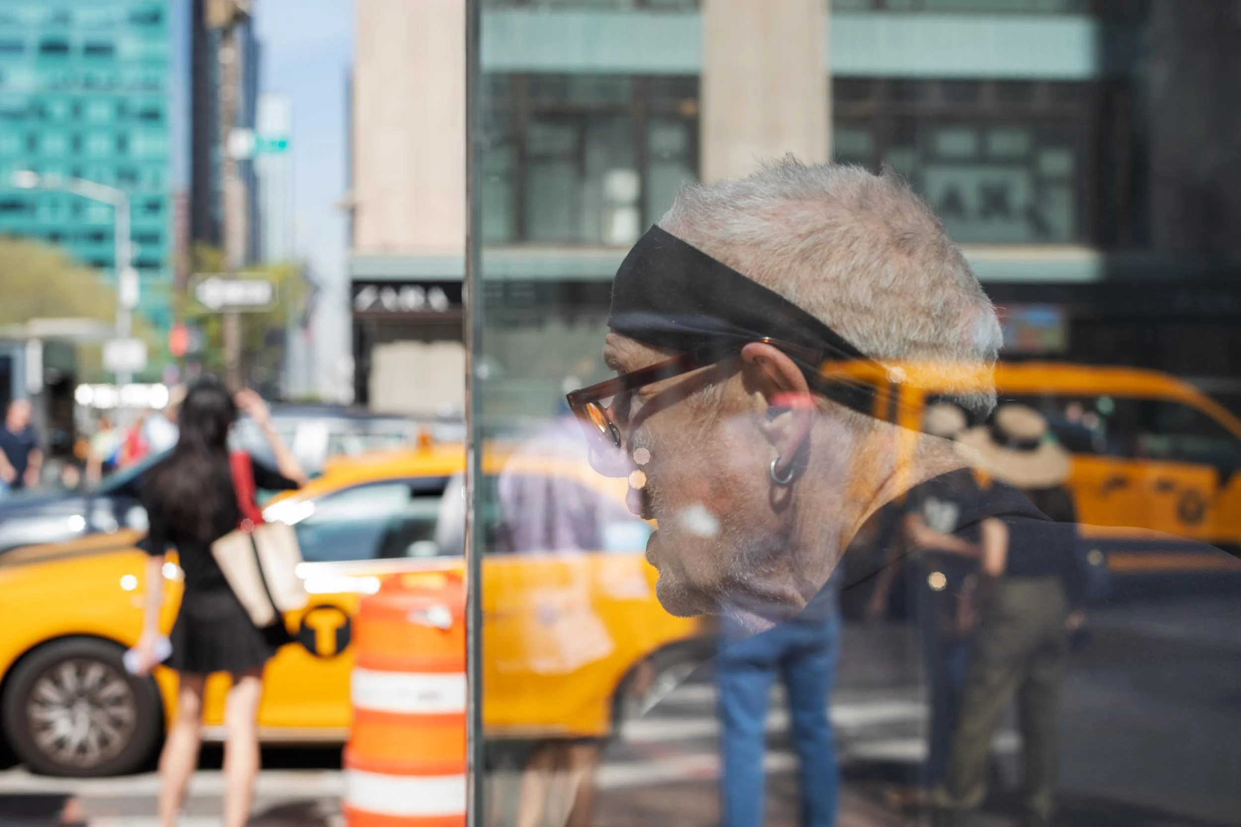 Eine grauhaarige Person hinter einer Glasscheibe, überlagert von Spiegelungen gelber New Yorker Taxis.