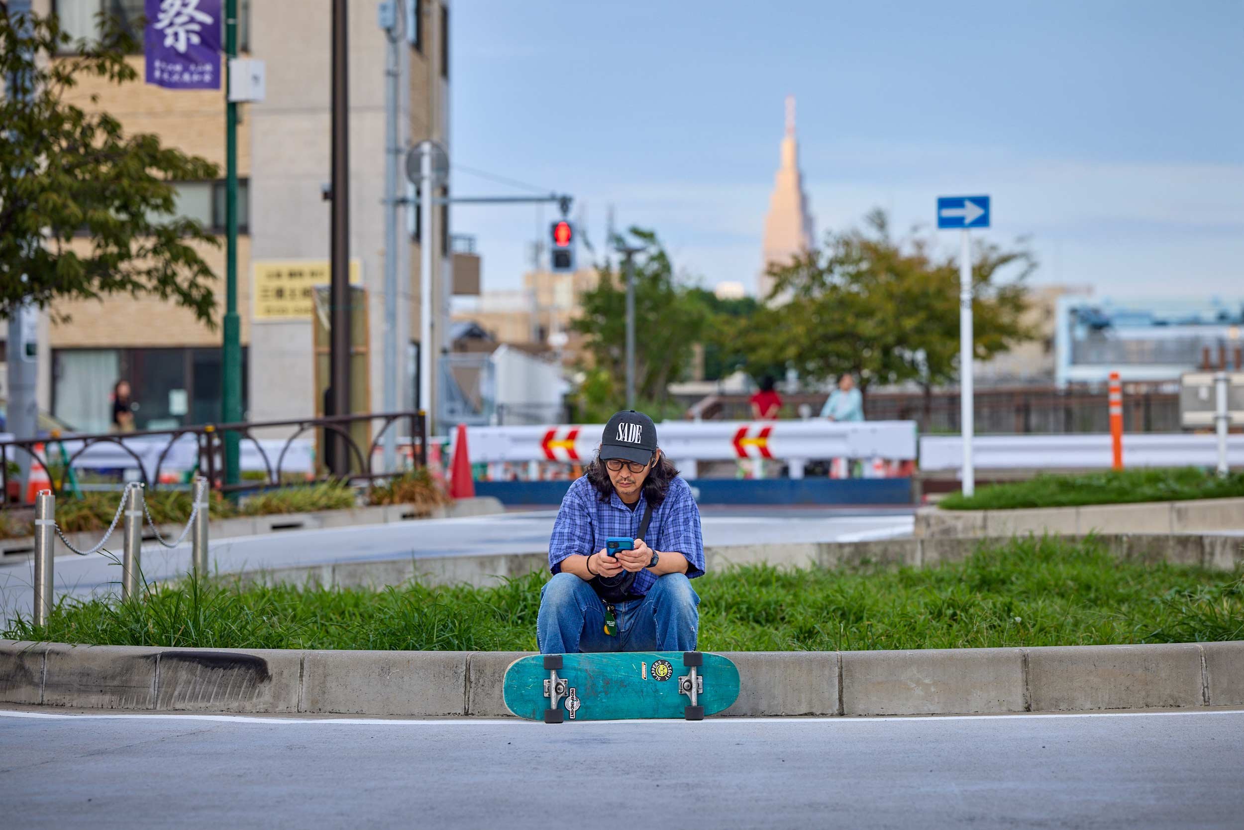 Mann sitzt auf einem Bordstein vor einer Grünfläche, ein Skateboard liegt vor ihm, er schaut auf sein Smartphone, im Hintergrund eine städtische Kreuzung.Tokio
