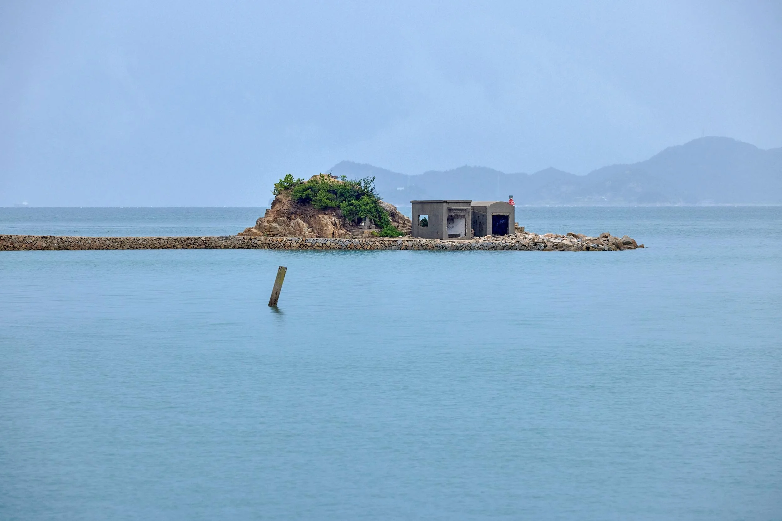 Kleine Insel mit Hafenmauer und Gebäude im ruhigen Meer vor einer Berglandschaft.
