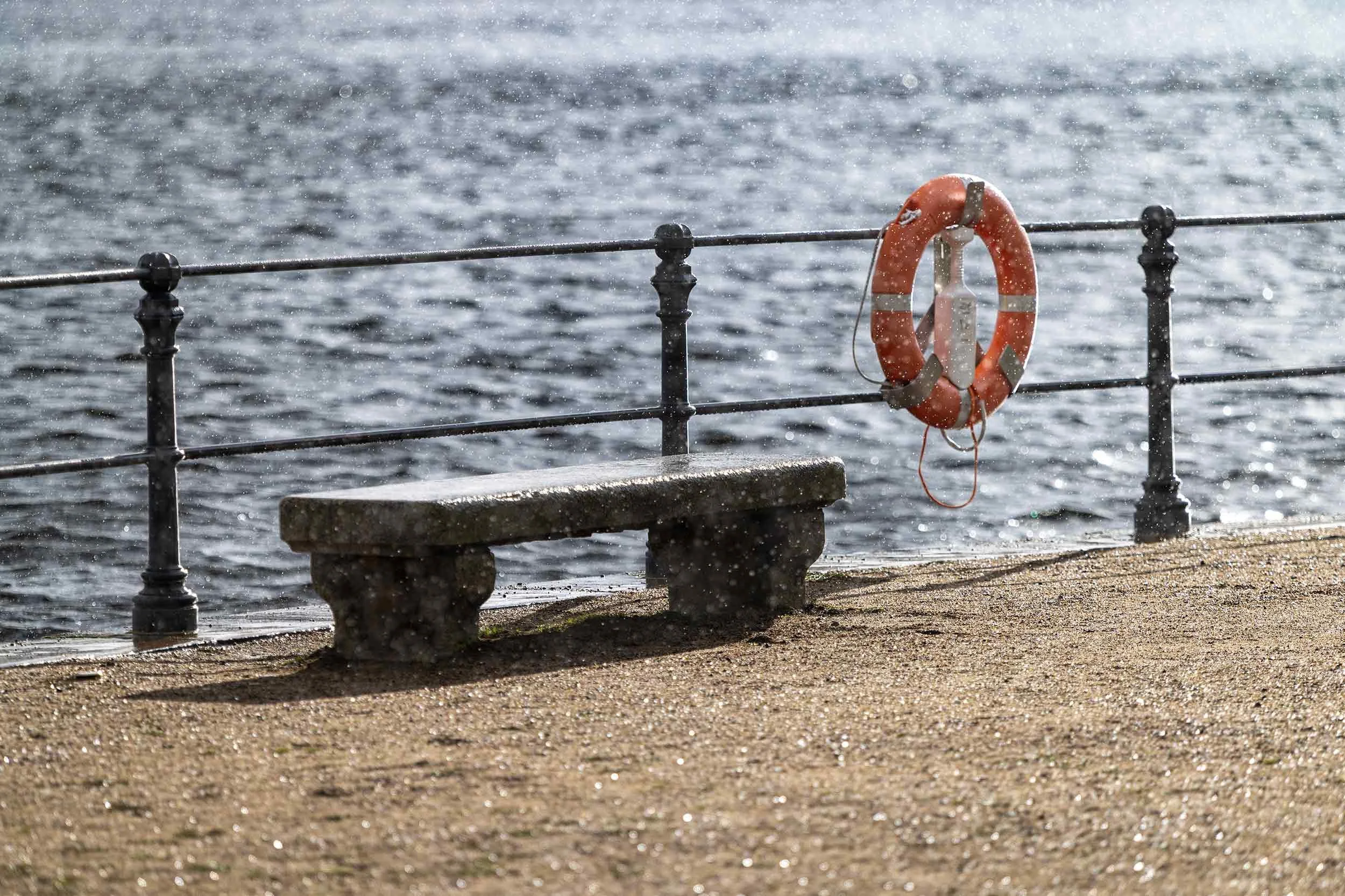 Eine leere Steinbank und ein orangefarbener Rettungsring stehen am Geländer unmittelbar an der Hamburger Alster. Vom Gegenlicht hervorgehobene Regentropfen schweben über dem Wasser und verwandeln die alltägliche Uferszene in einen atmosphärischen Mom