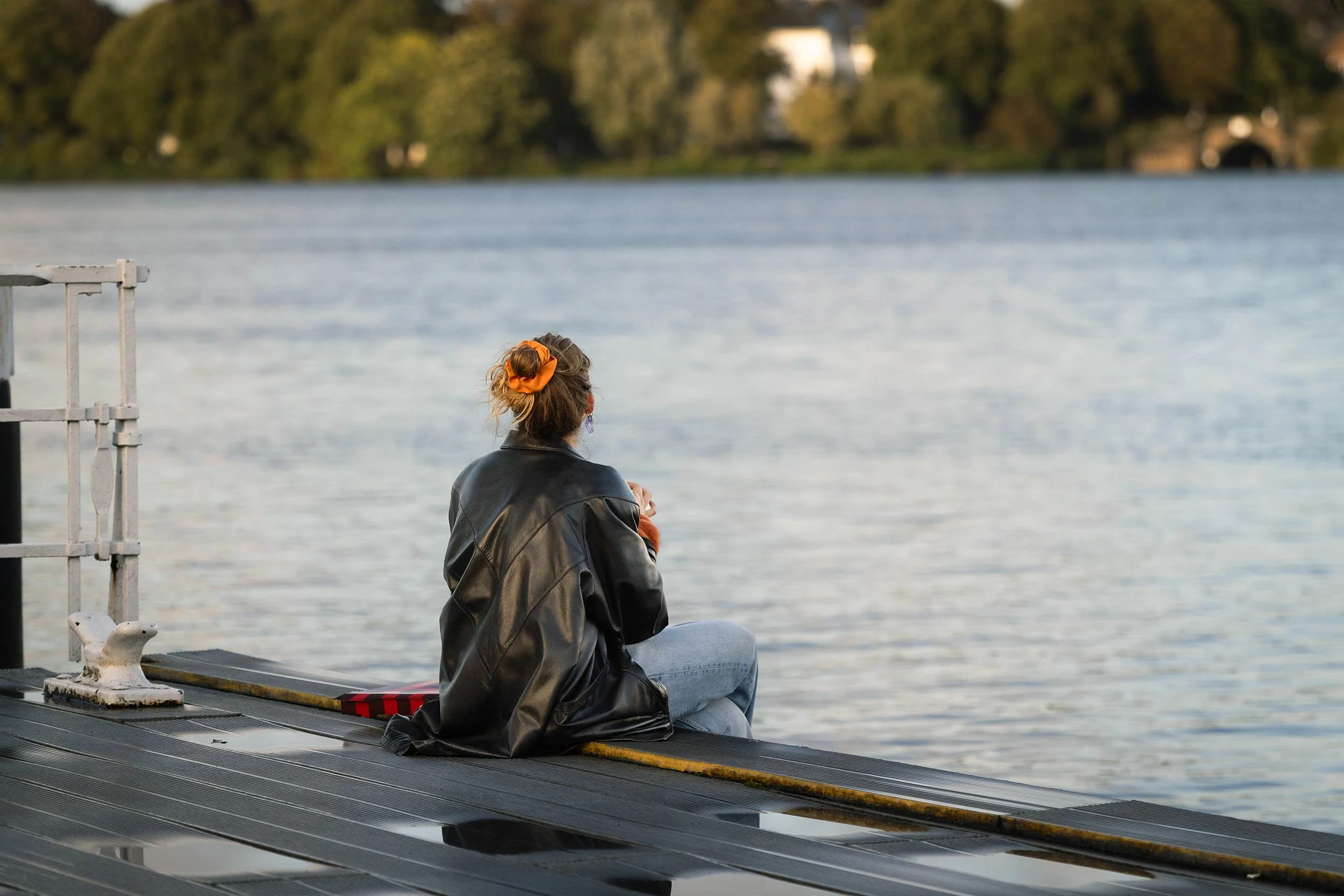 Eine junge Frau sitzt mit dem Rücken zur Kamera auf einem Anleger an der Hamburger Außenalster. Ihr orangefarbenes Haarband setzt einen kleinen Farbakzent zwischen schwarzer Lederjacke, hellem Wasser und der grünen Uferlandschaft im Hintergrund. Foto