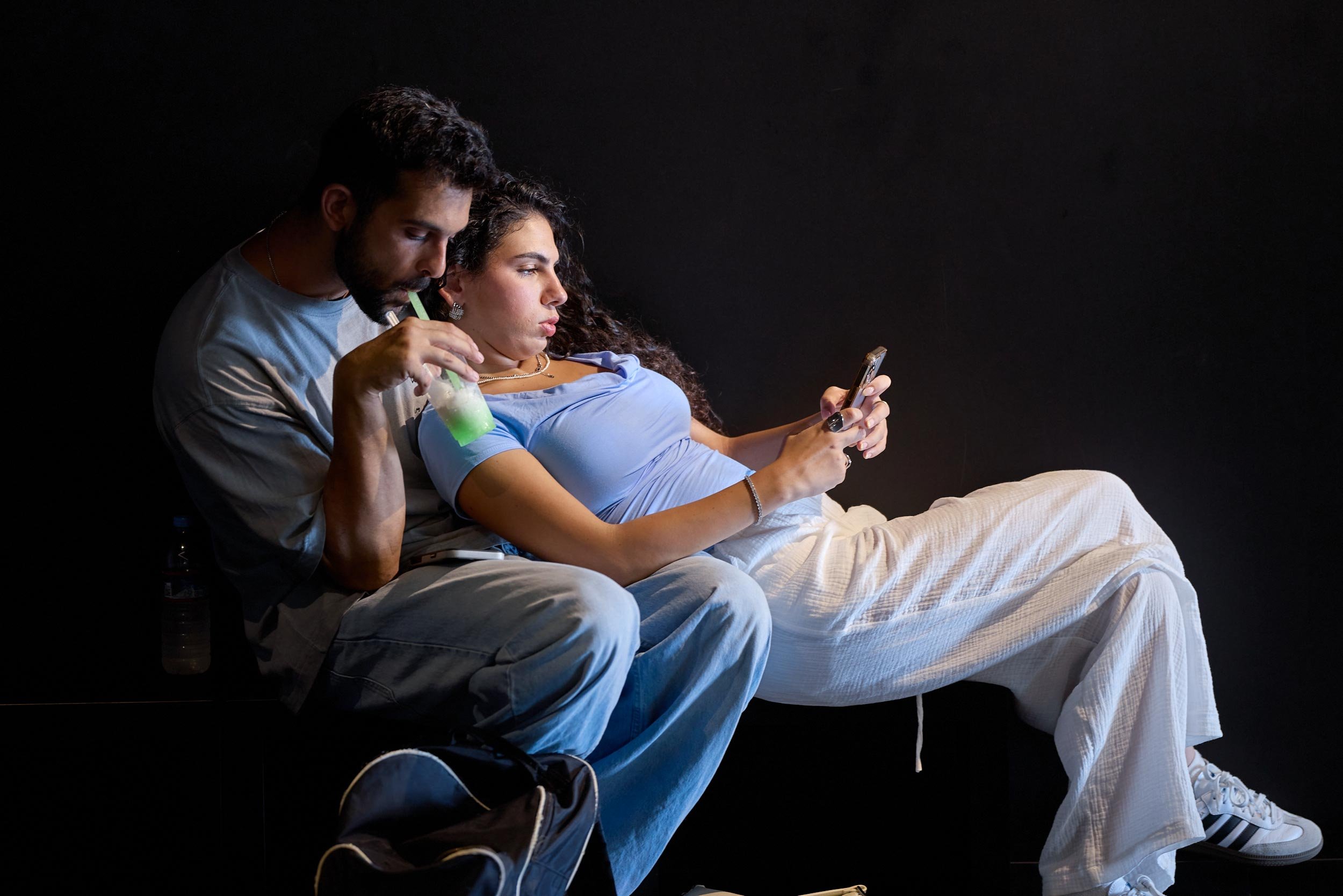 Paar sitzt nebeneinander in einem dunklen Raum, beide schauen auf ein Smartphone, eine Person hält ein Getränk.