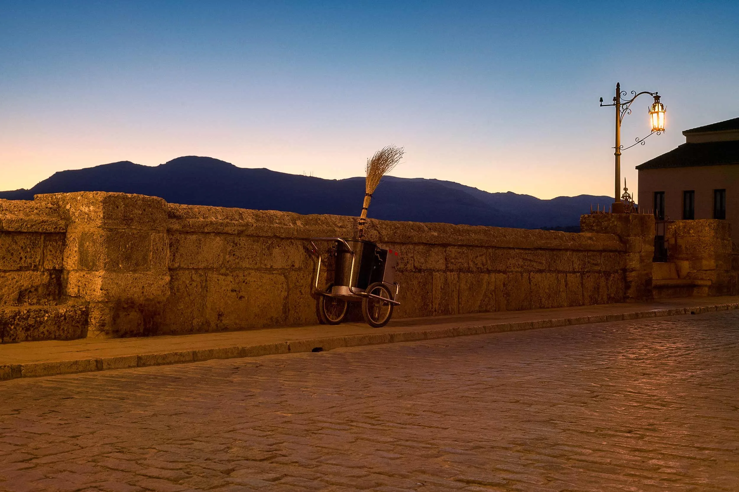 Eine stimmungsvolle Szene bei Dämmerung auf einer Brücke, beleuchtet von einer Straßenlaterne, während eine Karre mit einem Besen lehnt.