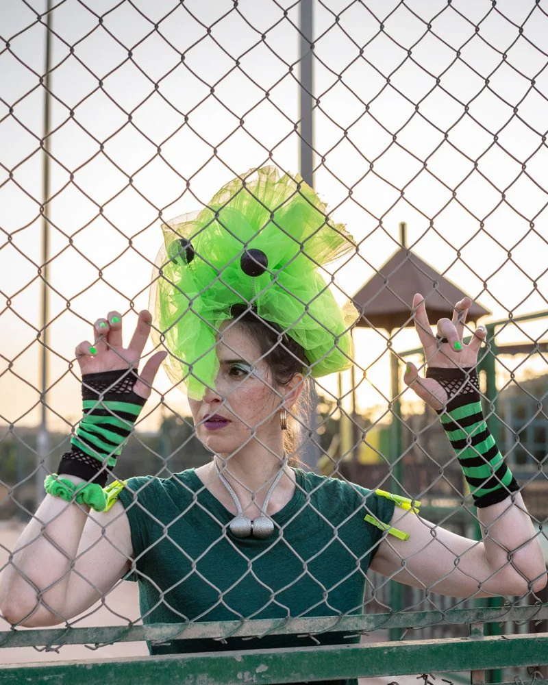 Frau mit neon-grünem Kopfschmuck steht hinter einem Maschendrahtzaun und hält den Zaun mit beiden Händen in Ibiza