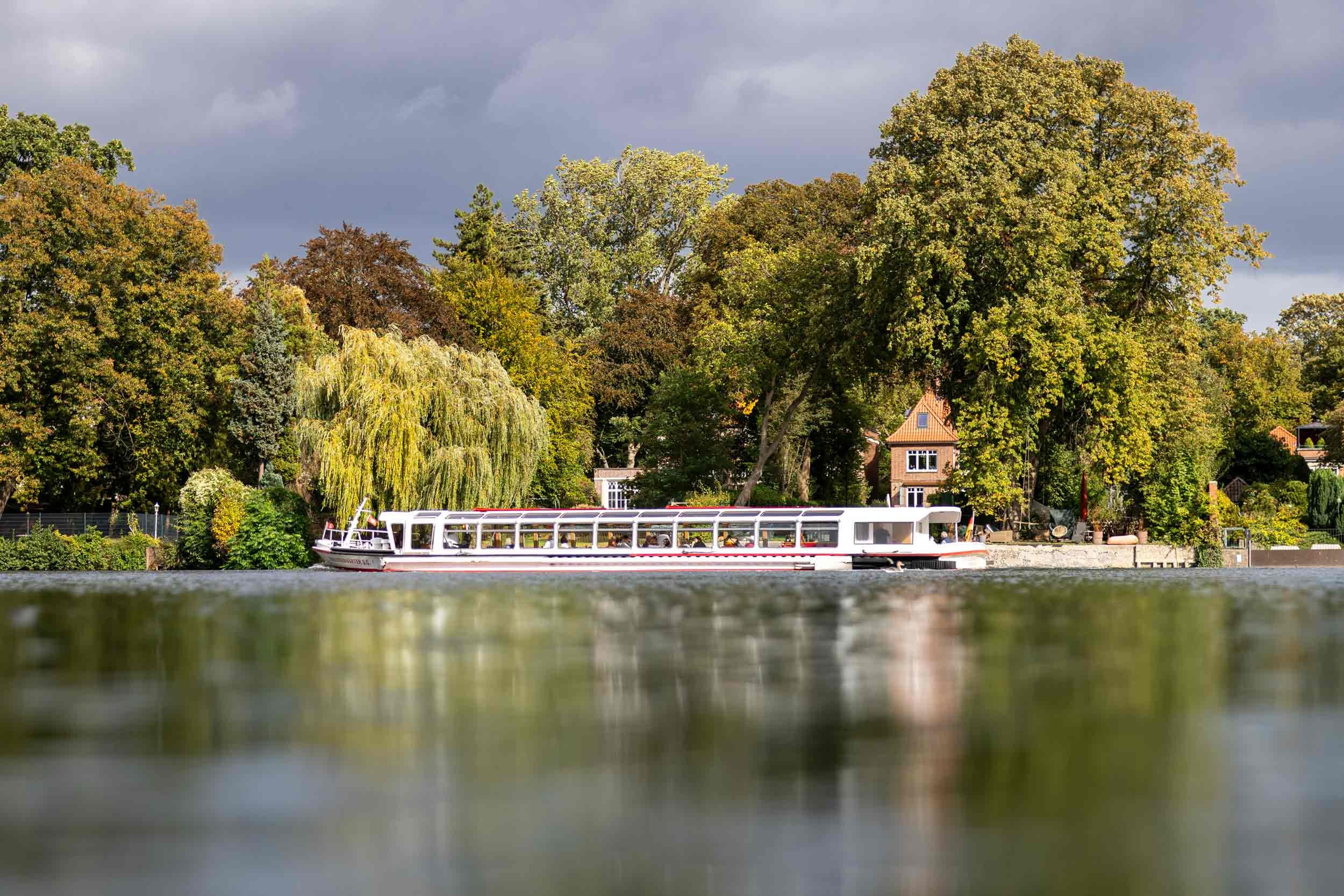 Das weiß-rote Alsterschiff Eilbek fährt über die Hamburger Außenalster. Bäume und Ufervegetation rahmen den Blick auf die dahinterliegende Stadtkulisse, während das bewegte Wasser und das warme Licht der Szene eine sommerliche Atmosphäre verleihen. F