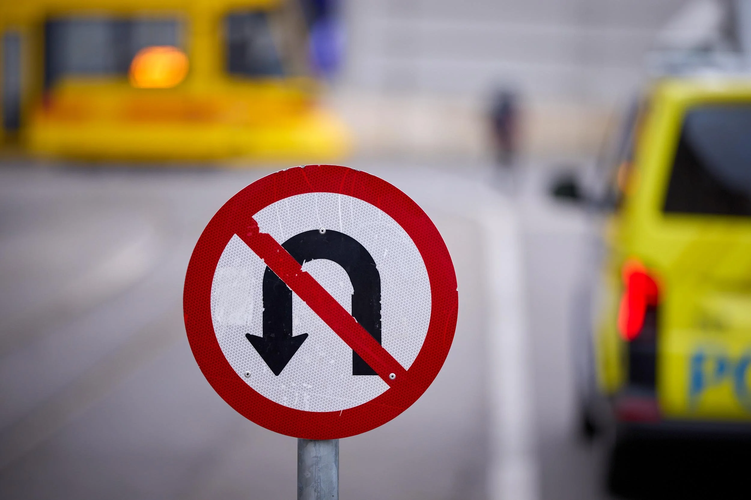 Rundes Verkehrsschild mit durchgestrichenem Wendepfeil am Straßenrand, Fahrzeuge im Hintergrund.
