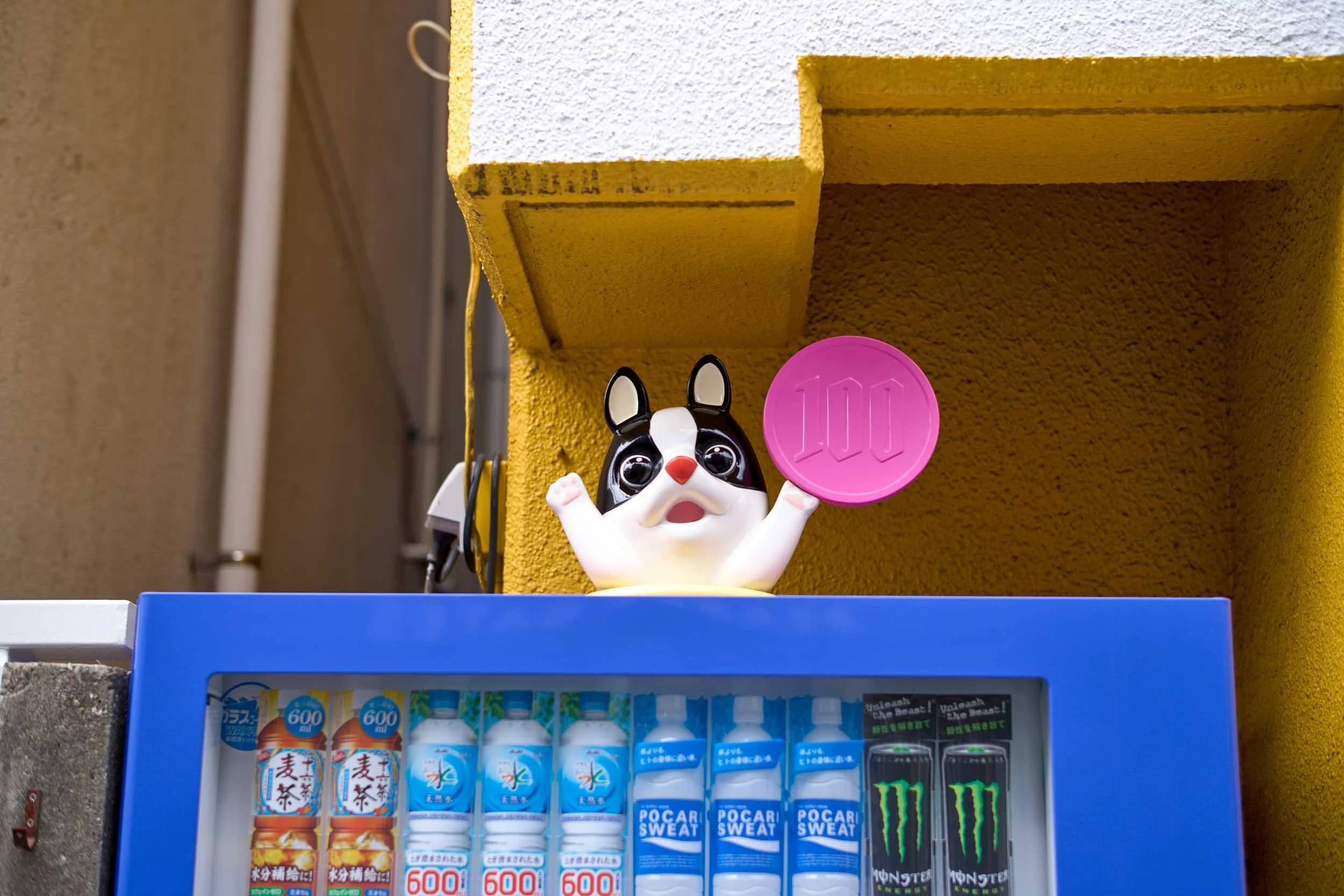 Hundefigur mit erhobenen Pfoten steht auf einem Getränkeautomaten vor einer gelben Hauswand. Tokio
