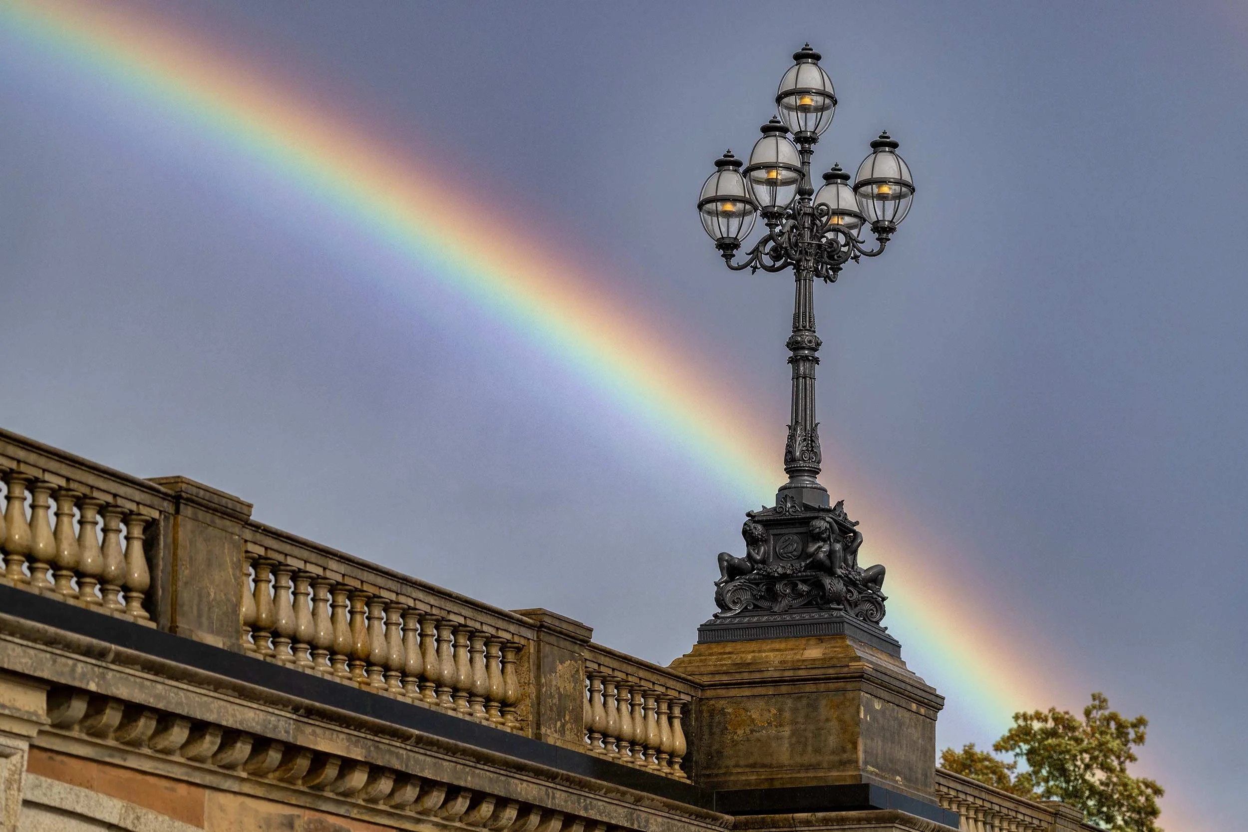 Ein kräftiger Regenbogen zieht sich diagonal über den dunklen Himmel hinter einer historischen Laterne der Lombardsbrücke in Hamburg. Die ornamentierte Leuchte und die steinerne Balustrade bilden einen markanten Kontrast zu den intensiven Farben des 