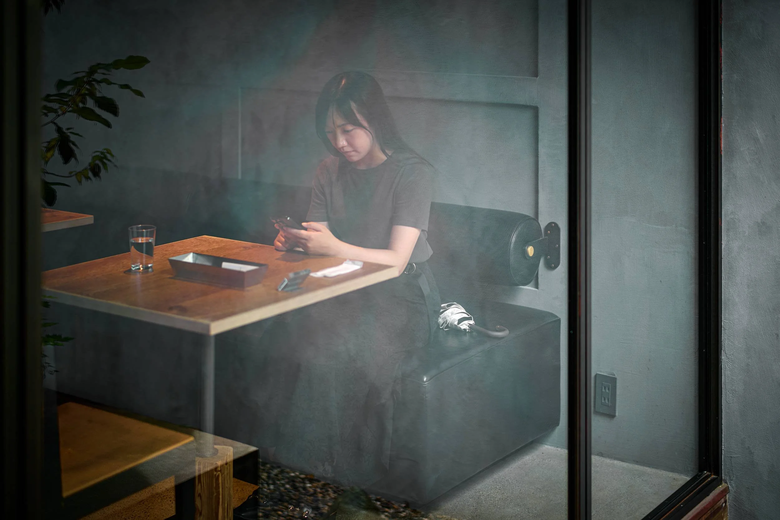 Frau sitzt in einem Café auf einer Bank und schaut auf ihr Smartphone, fotografiert durch eine Fensterscheibe.